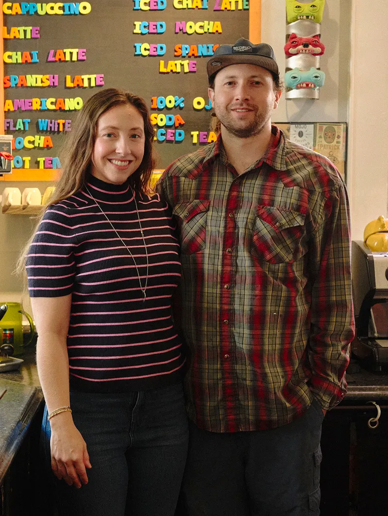 Smiling man and woman standing together in a cafe with a colorful menu board behind them.