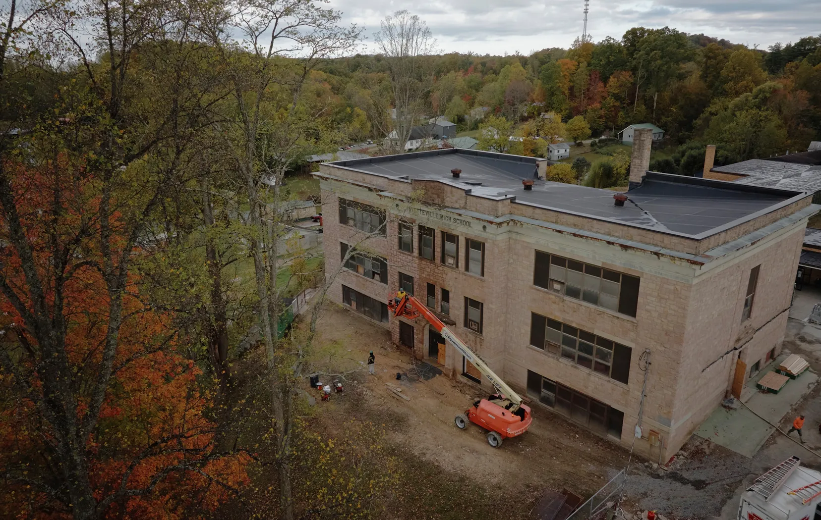 Aerial view of an old school building undergoing exterior renovation with a cherry picker and workers visible.
