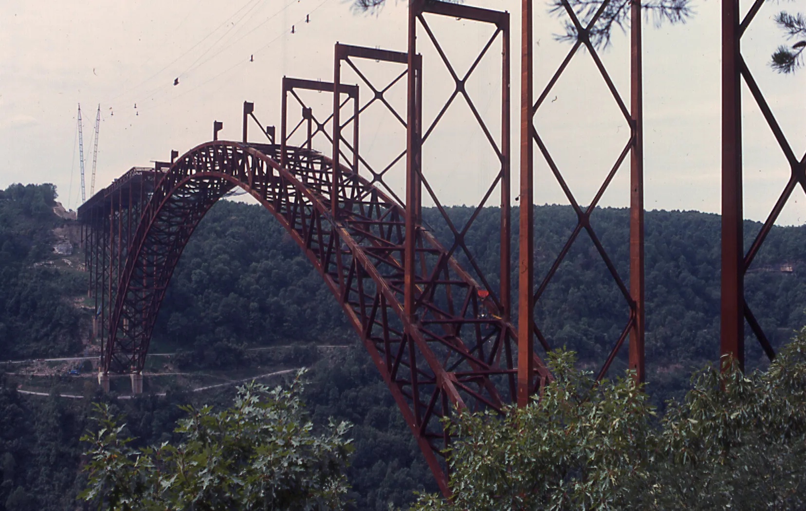 Rust-colored steel arch bridge under construction spanning a deep forested valley.