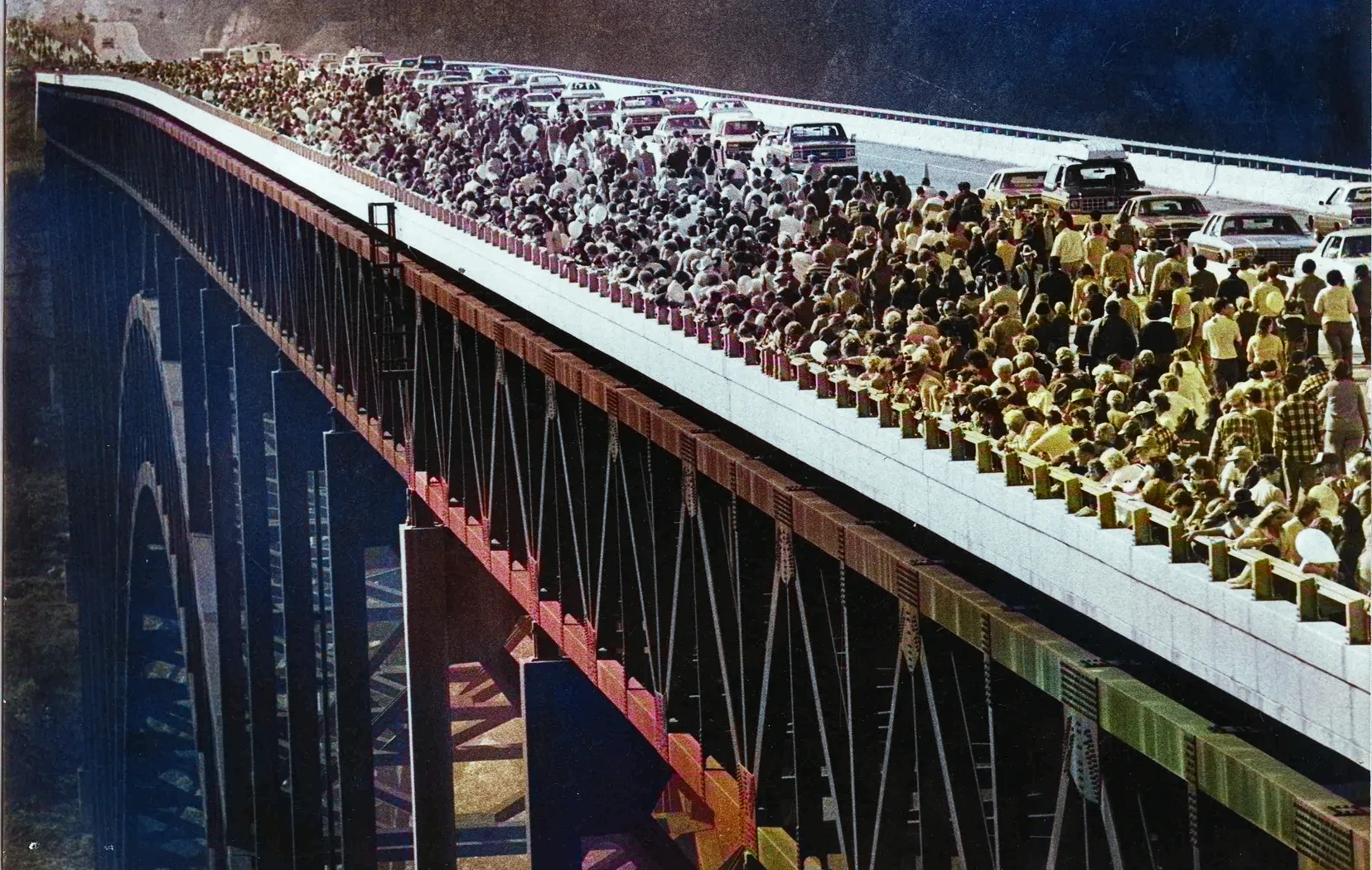 Large crowd of people walking across a high bridge, with cars stopped behind them.