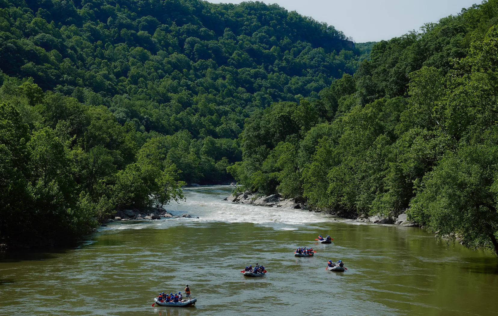 Several groups of people rafting on a river surrounded by dense green forest and hills.