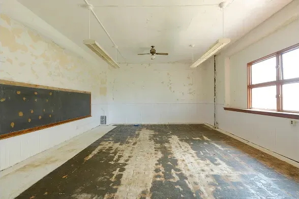 Empty, worn classroom with peeling walls, old blackboard, and large window letting in daylight.
