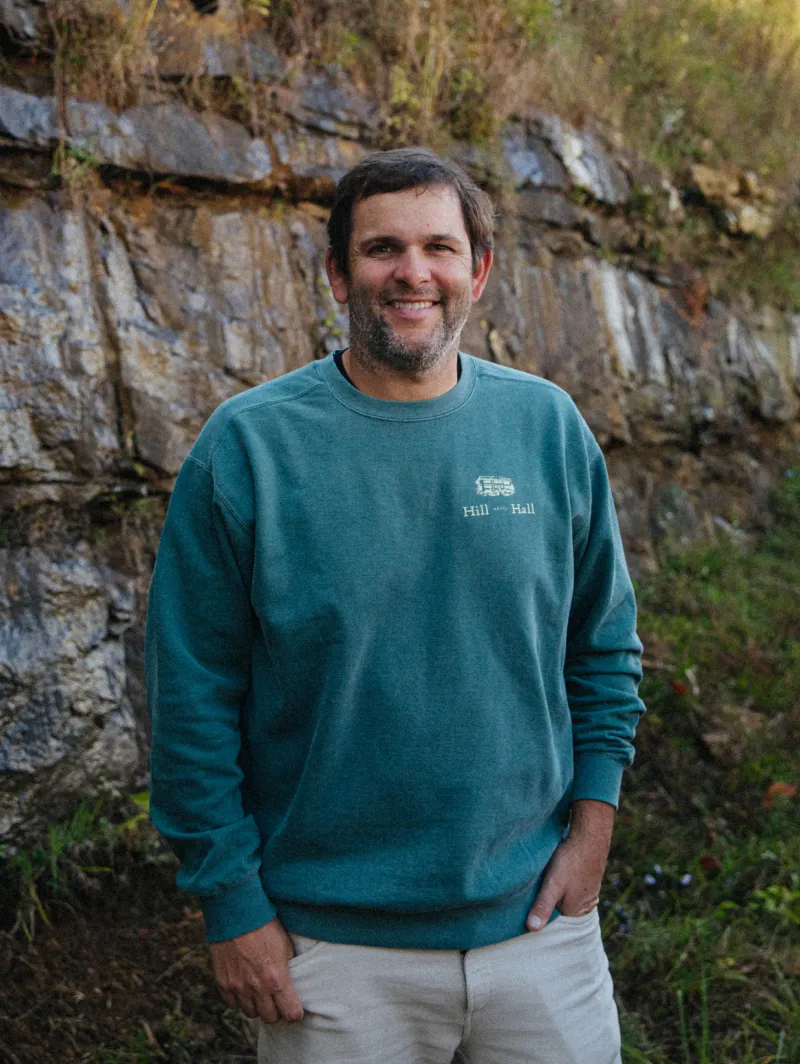 Smiling man with short dark hair and beard wearing a teal sweatshirt standing outdoors in front of rocky terrain.