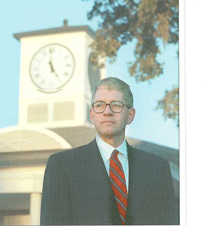 Man in glasses and dark suit with red striped tie standing outdoors in front of a clock tower showing 11:30.