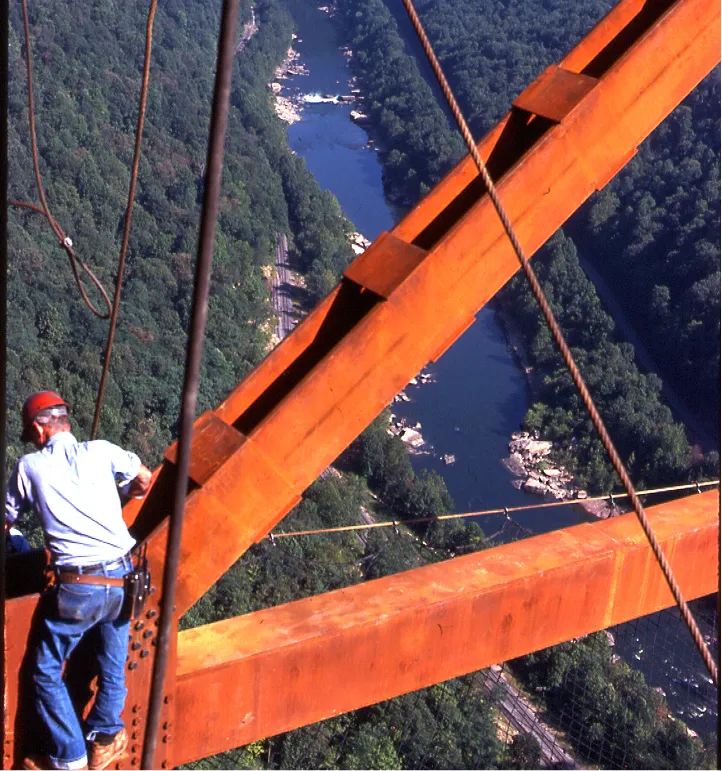 Worker wearing a red helmet and blue jeans climbing orange steel beams above a forested river valley.