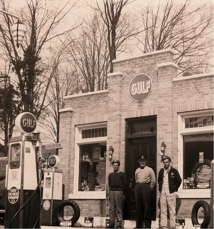 Three men standing in front of a vintage Gulf gas station with two fuel pumps and tire displays.