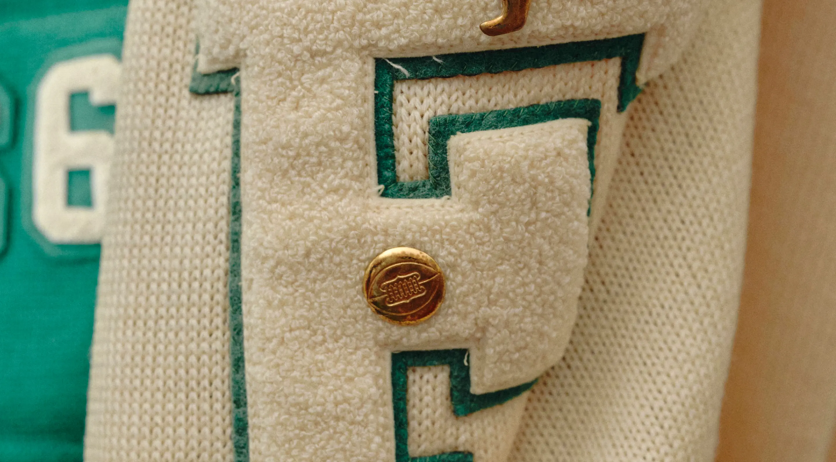 Close-up of a cream-colored textured letter patch with green edging and a gold button featuring a stitched football design.