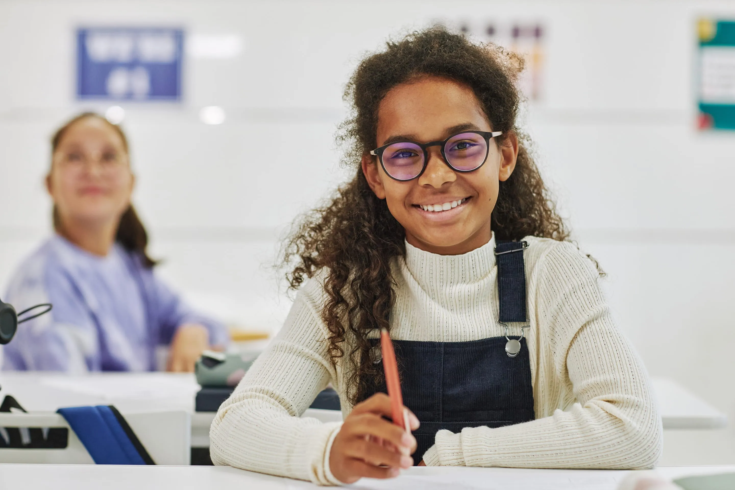 Smiling girl with curly hair and glasses sitting at a desk holding a pencil in a classroom.