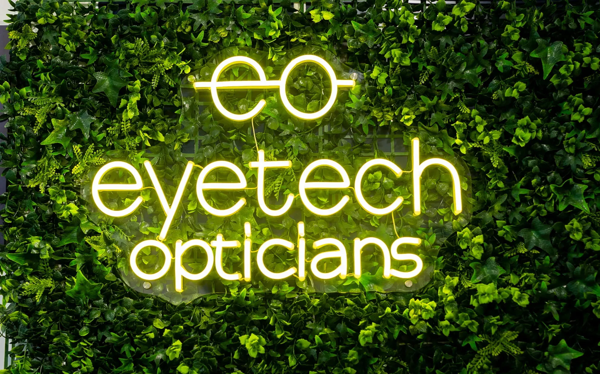 Yellow neon sign reading 'eye tech opticians' with eyeglasses icon on a green leafy background.