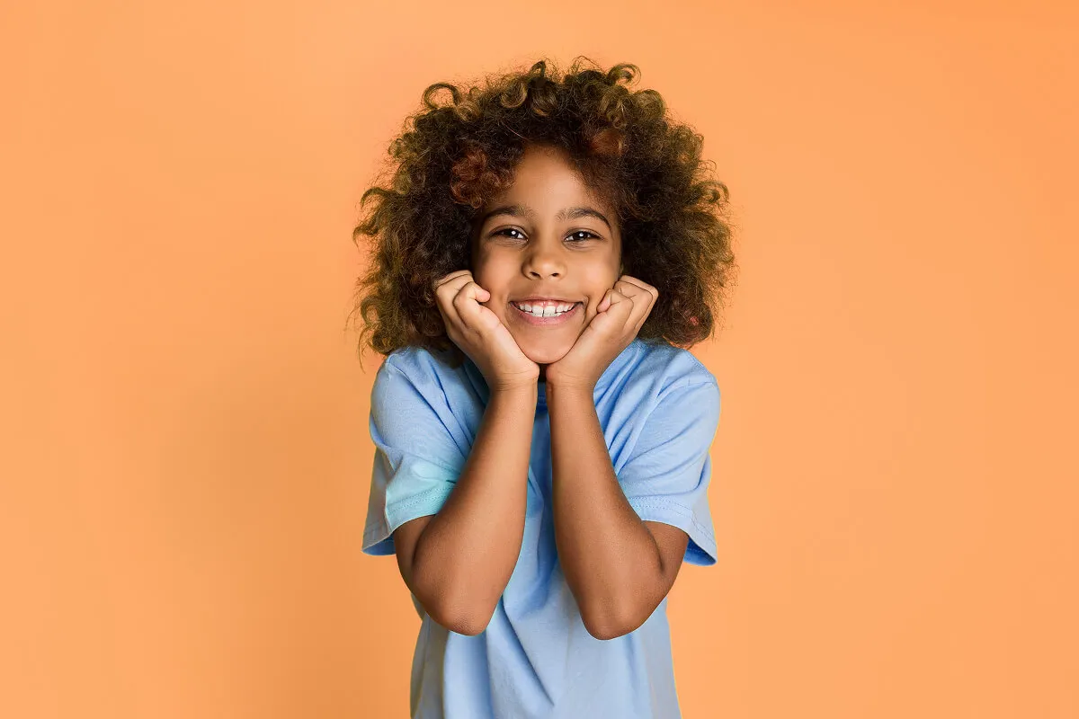 Smiling child with curly hair resting chin on hands against an orange background.