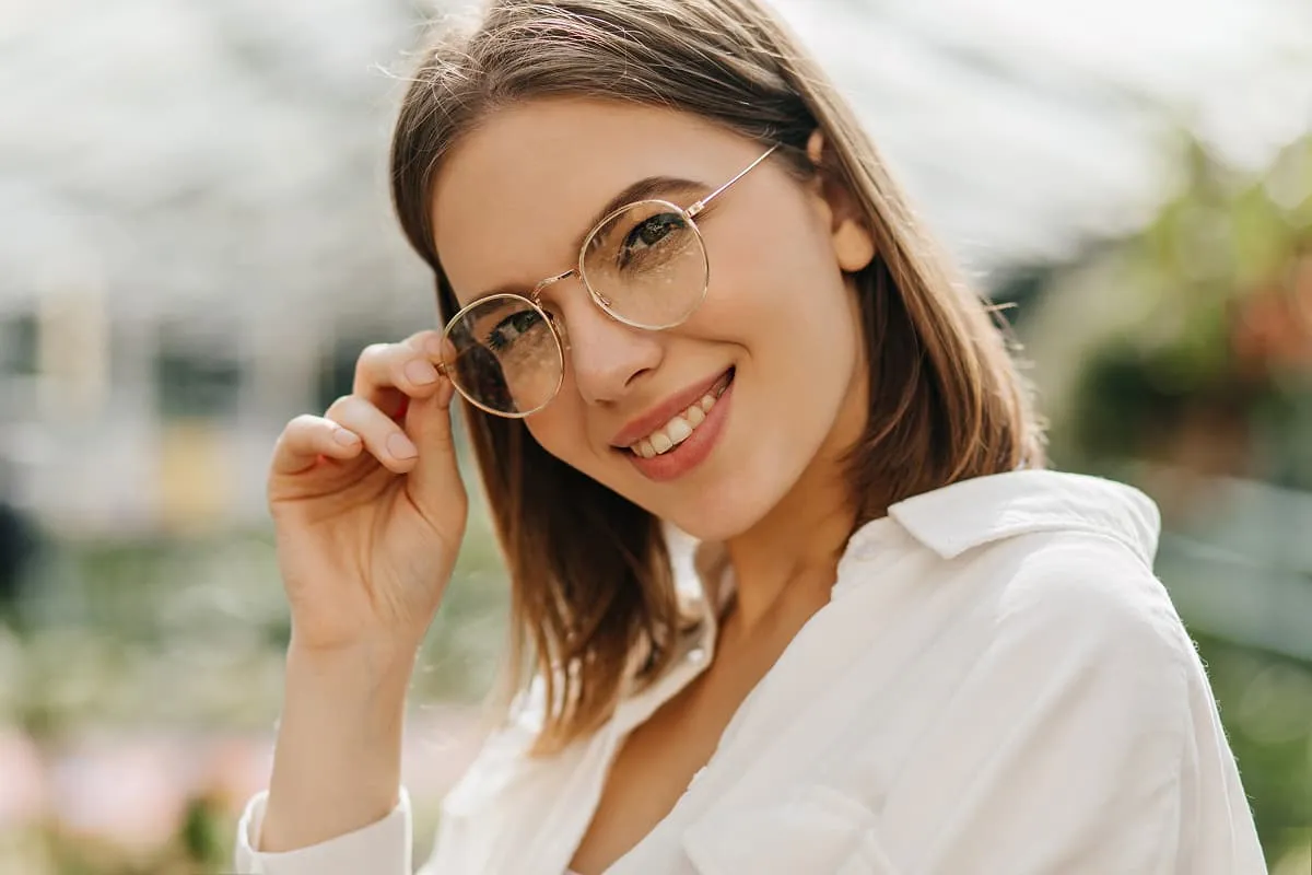 Smiling woman wearing round elegant eyeglasses and a white shirt, holding the frame with one hand.