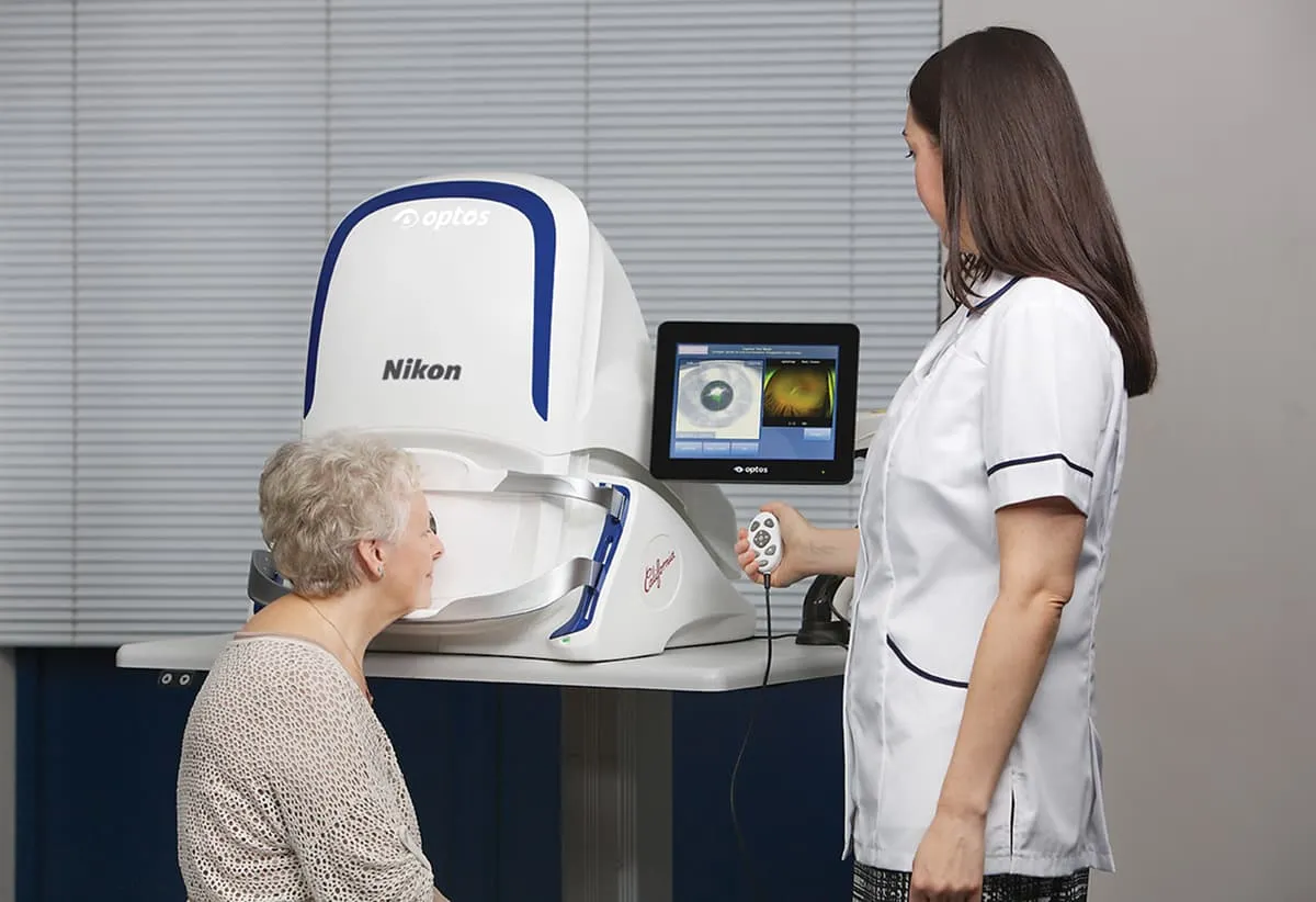 Older woman having an eye scan using a Nikon Optos machine operated by a healthcare professional.