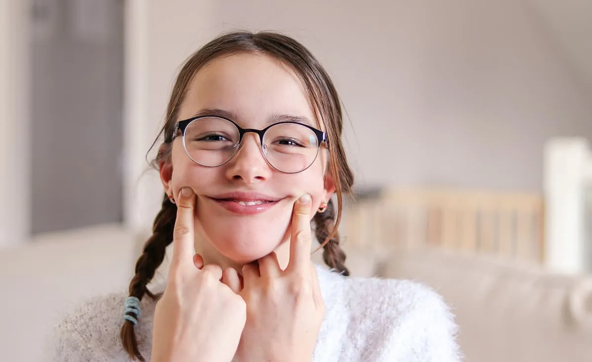 Young girl with glasses smiling and using her fingers to stretch her smile wider.