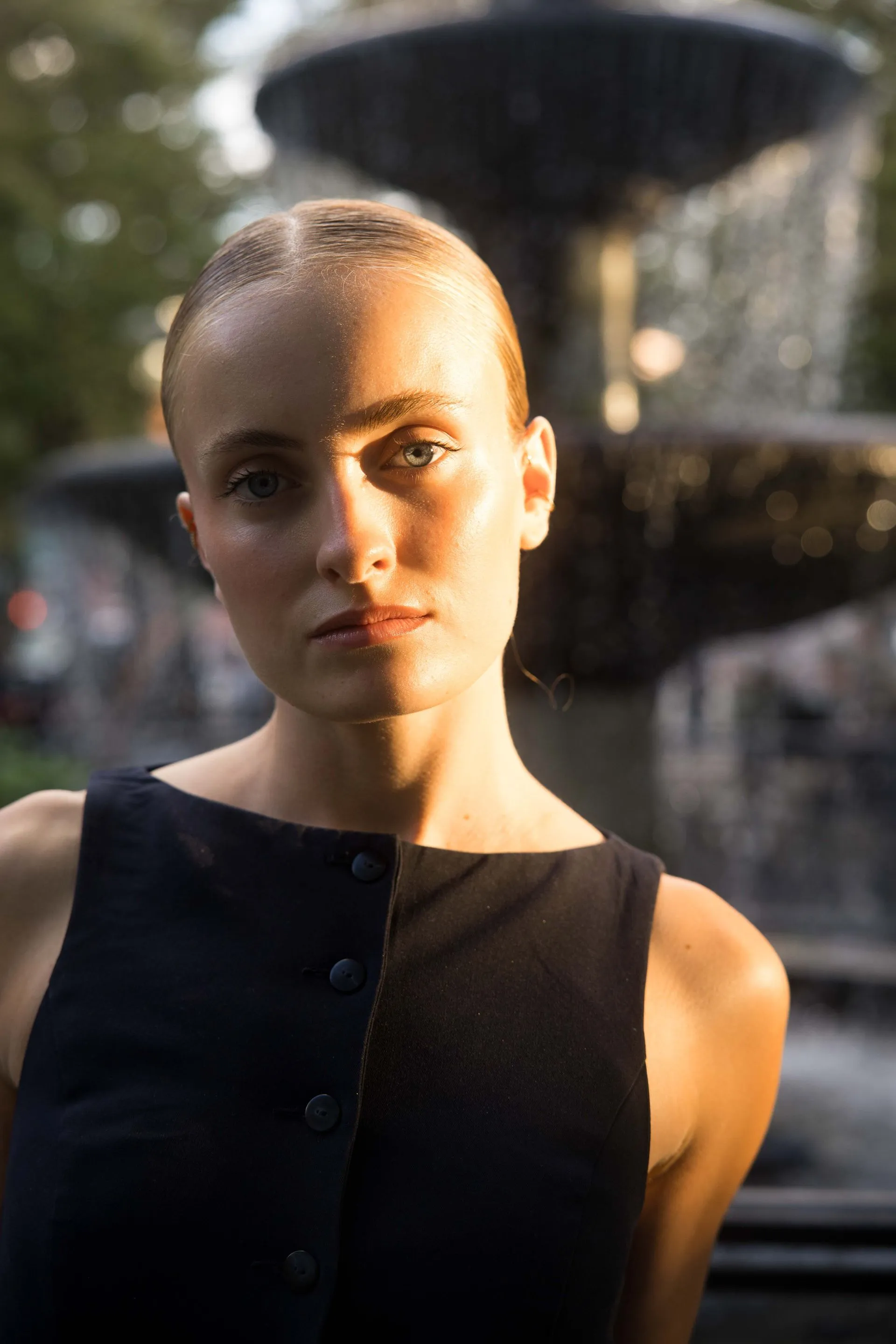 Woman in dark sleeveless top, lit by sunlight, in front of a fountain