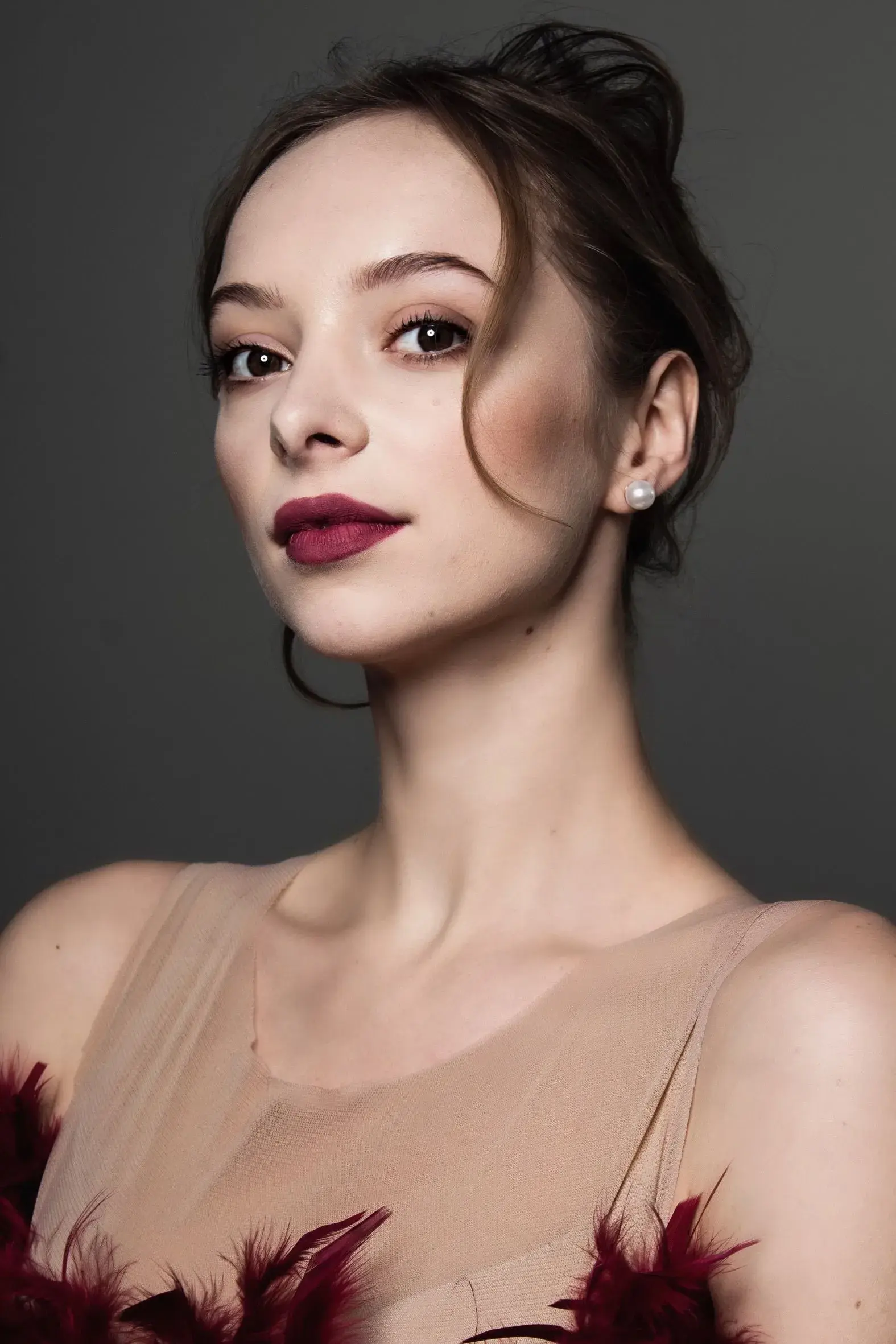 Woman with updo, dark red lipstick, wearing a beige sheer top with red feathers, against a gray backdrop