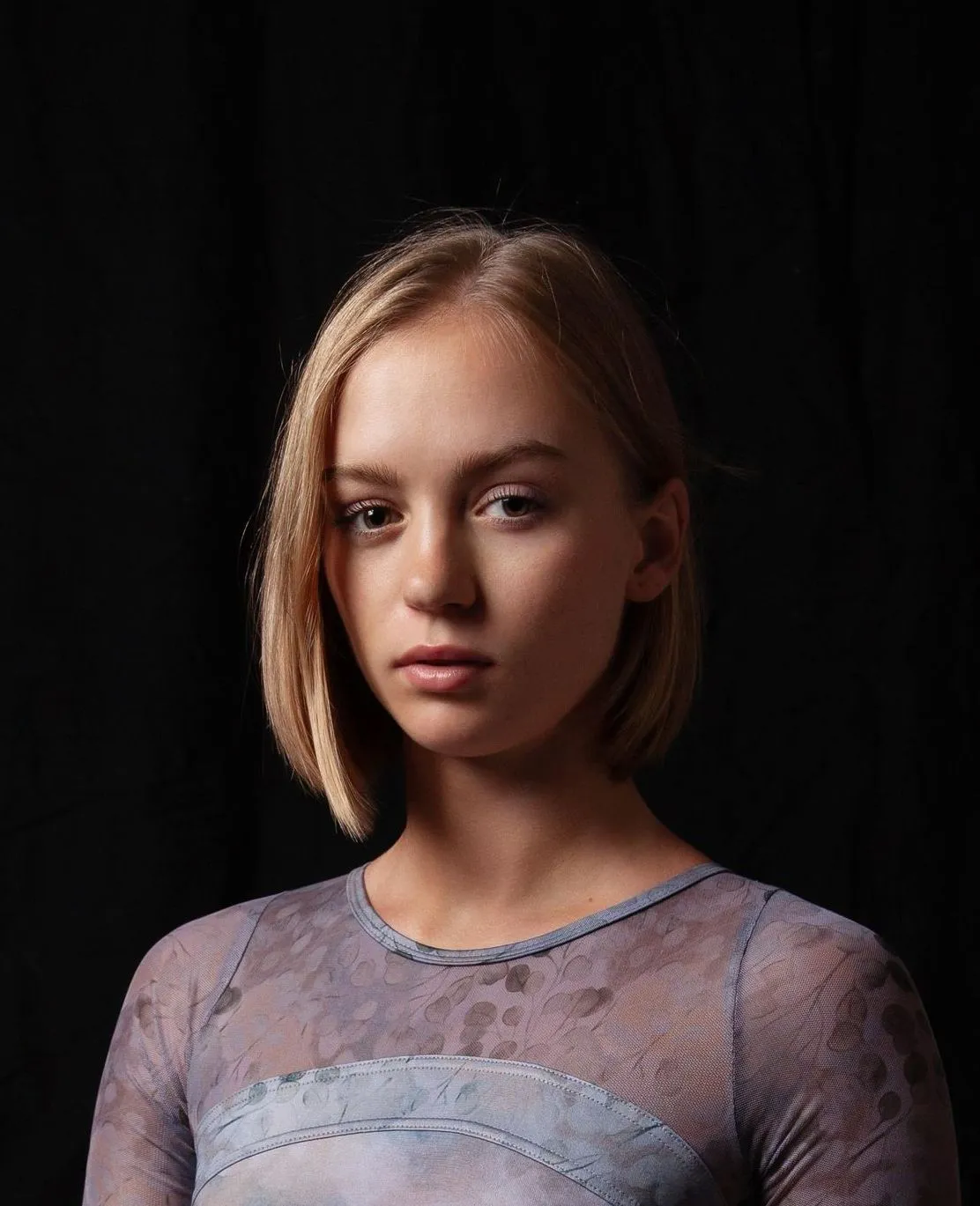 Woman with short blonde hair and a serious expression, wearing a patterned blue top against a black backdrop