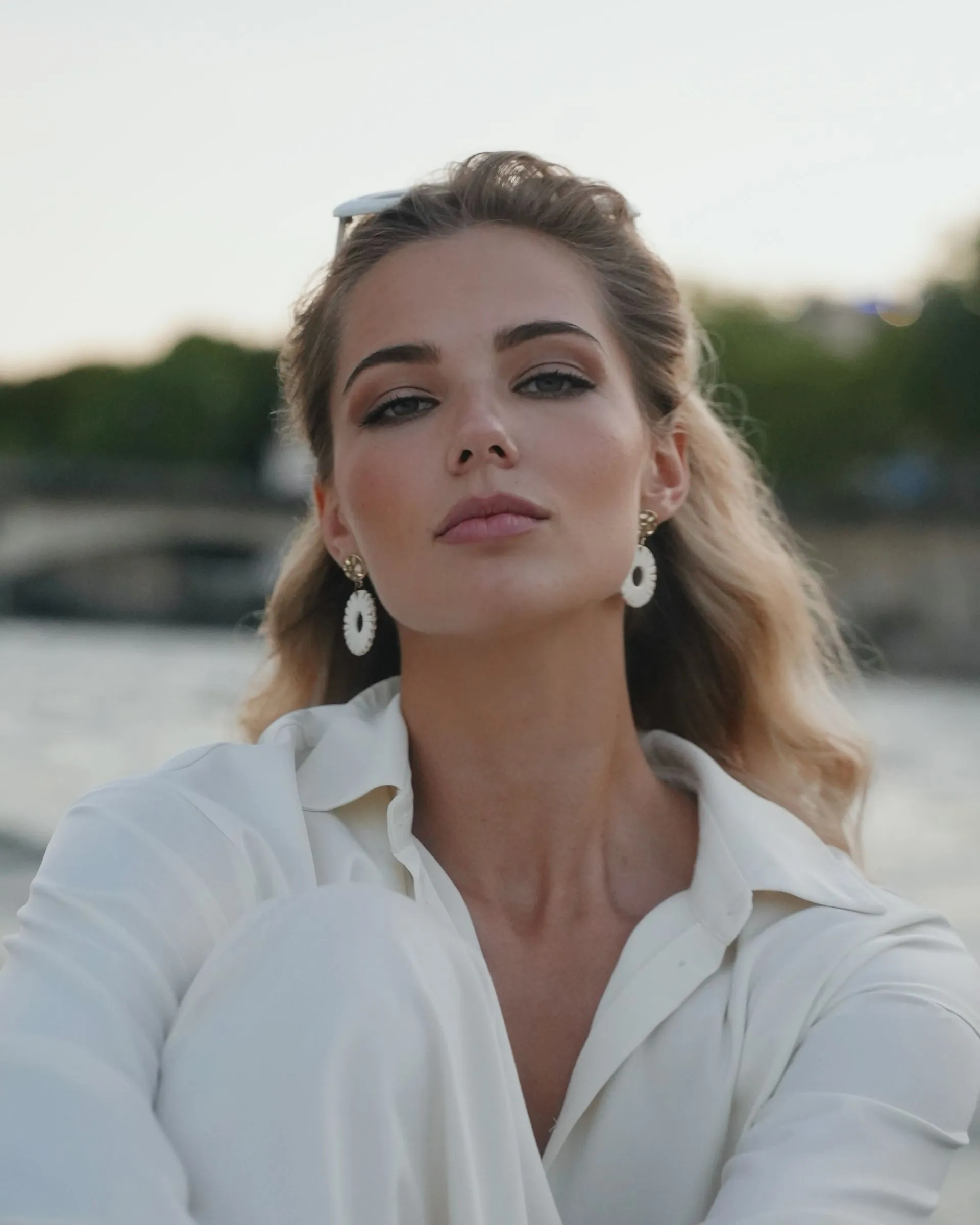 Woman with blonde hair wearing white outfit and earrings, looking at camera outdoors