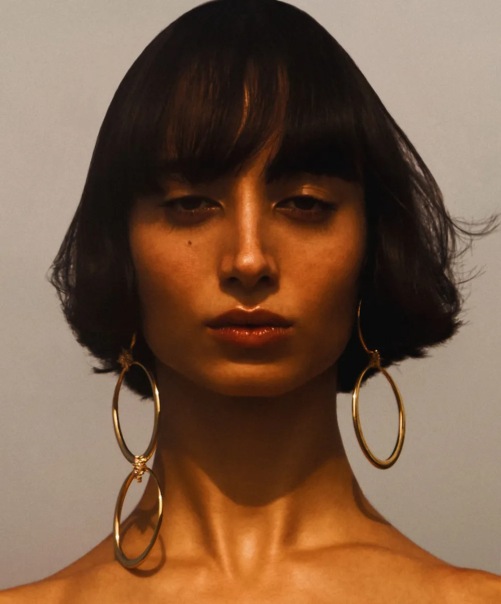 Woman with short dark hair and bangs, wearing large gold hoop earrings, looking directly at the viewer