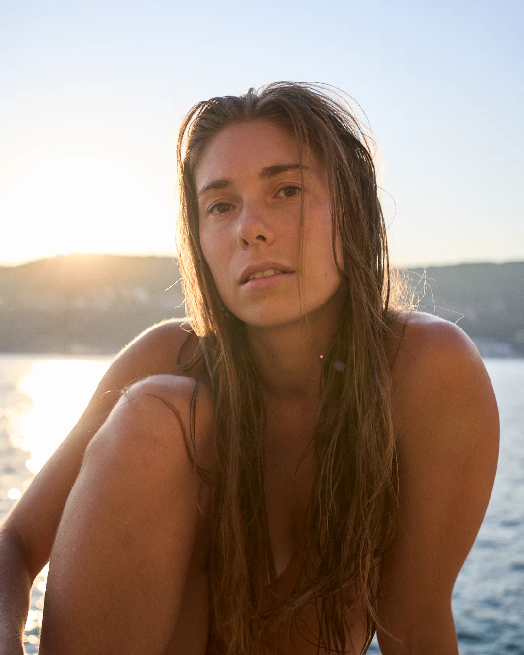 Woman with wet hair sits near water, looking towards the camera, sun sets in the background