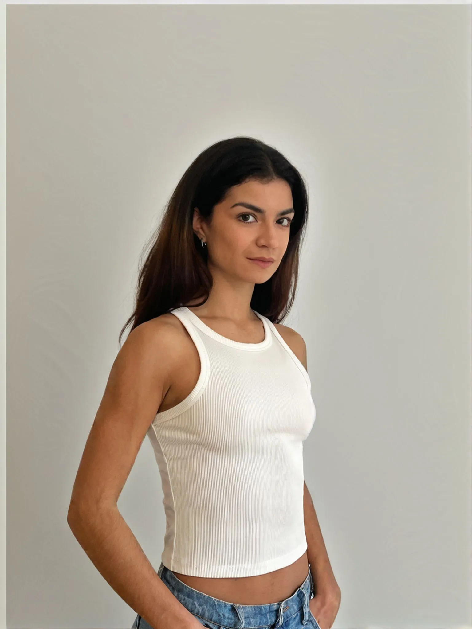 Woman in white ribbed tank top and jeans, hands in pockets, looking at the camera