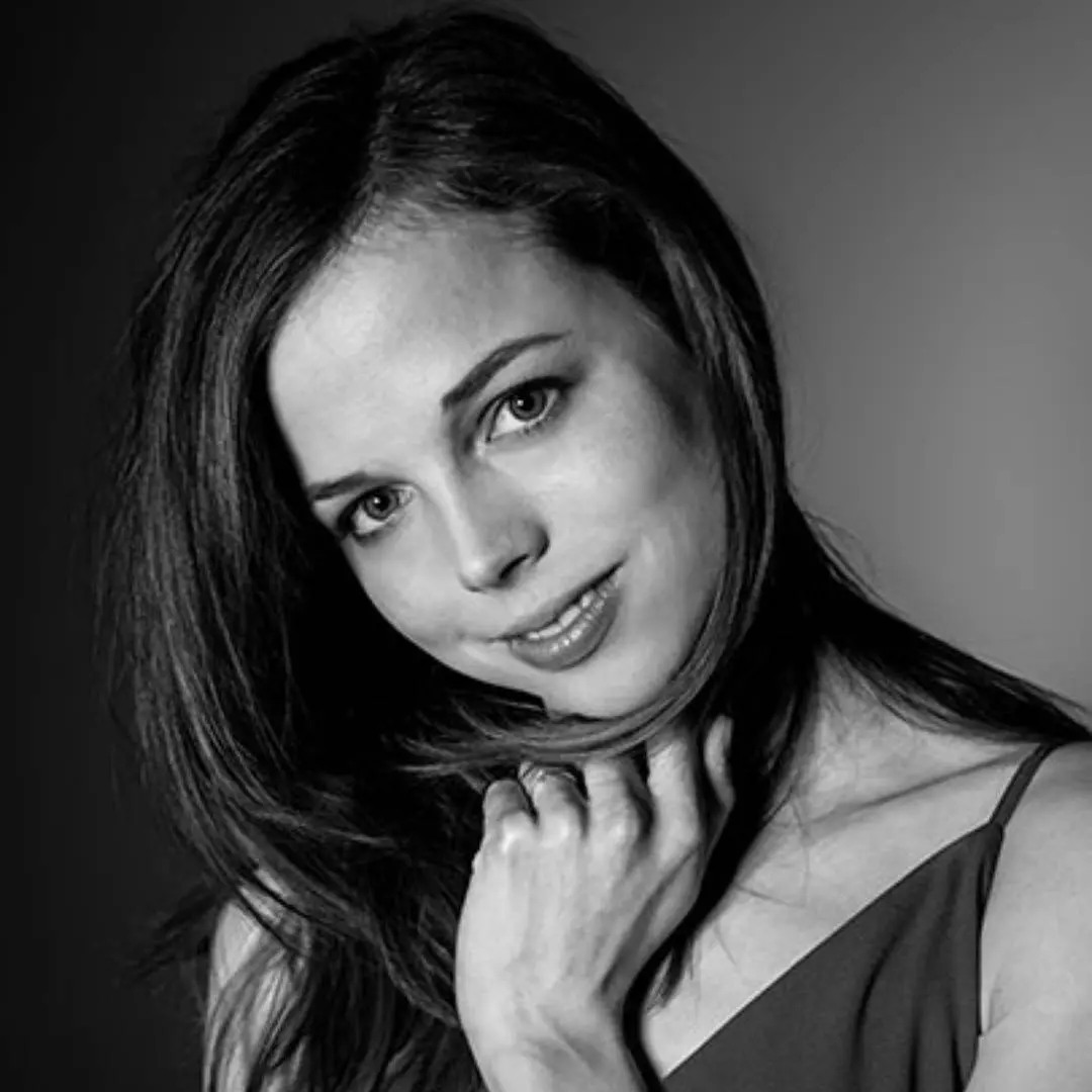 A woman in a dress touches her face with her hand and smiles