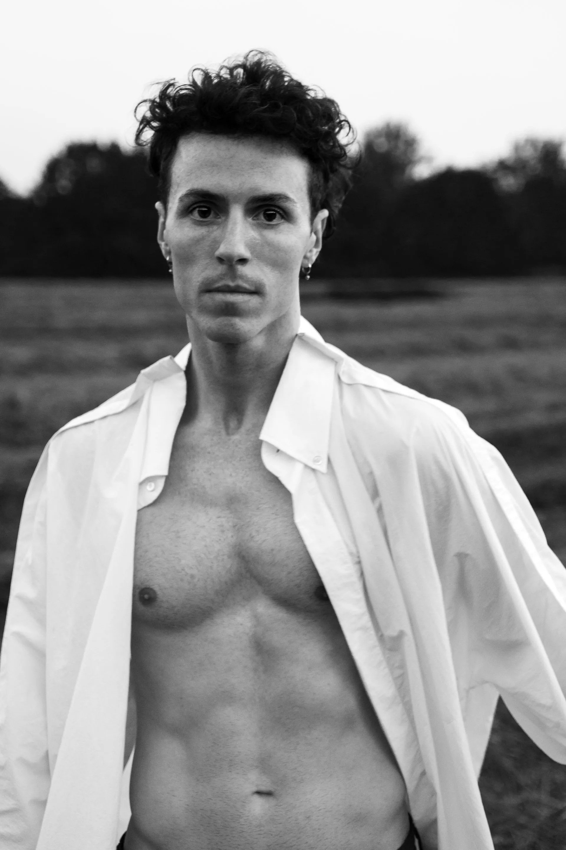 Man, shirtless under an open white shirt, stands in a field