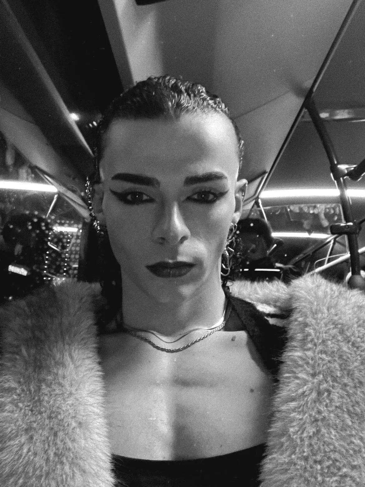 Black-and-white close-up portrait of a man with slicked-back hair, bold eye makeup, and earrings, wearing a fur coat and looking directly at the camera inside a dimly lit vehicle