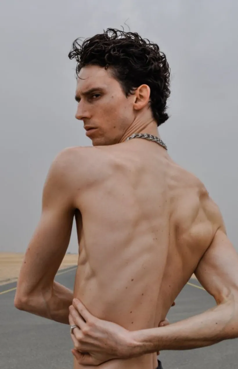 Man with curly hair, shirtless, turning to look over his shoulder; chain necklace, gray sky, open road