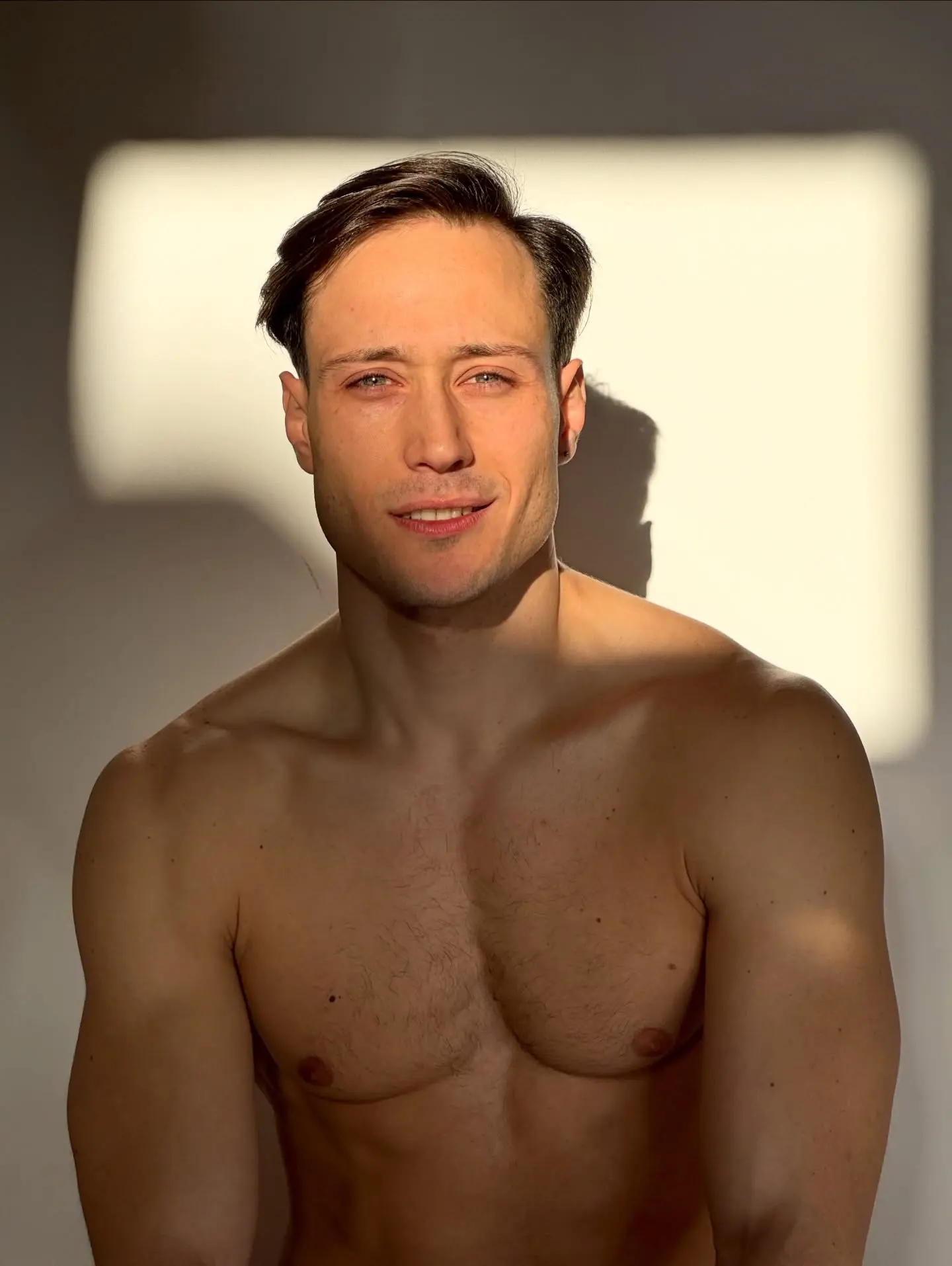Portrait of a shirtless man with short dark hair, standing against a light wall with warm sunlight casting shadows across his face and upper body, looking at the camera with a slight smile