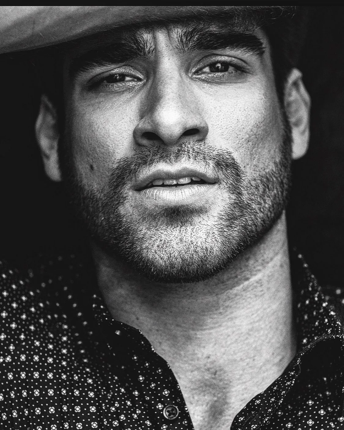 Man with a beard, wearing patterned shirt, hand on forehead, looking toward the viewer