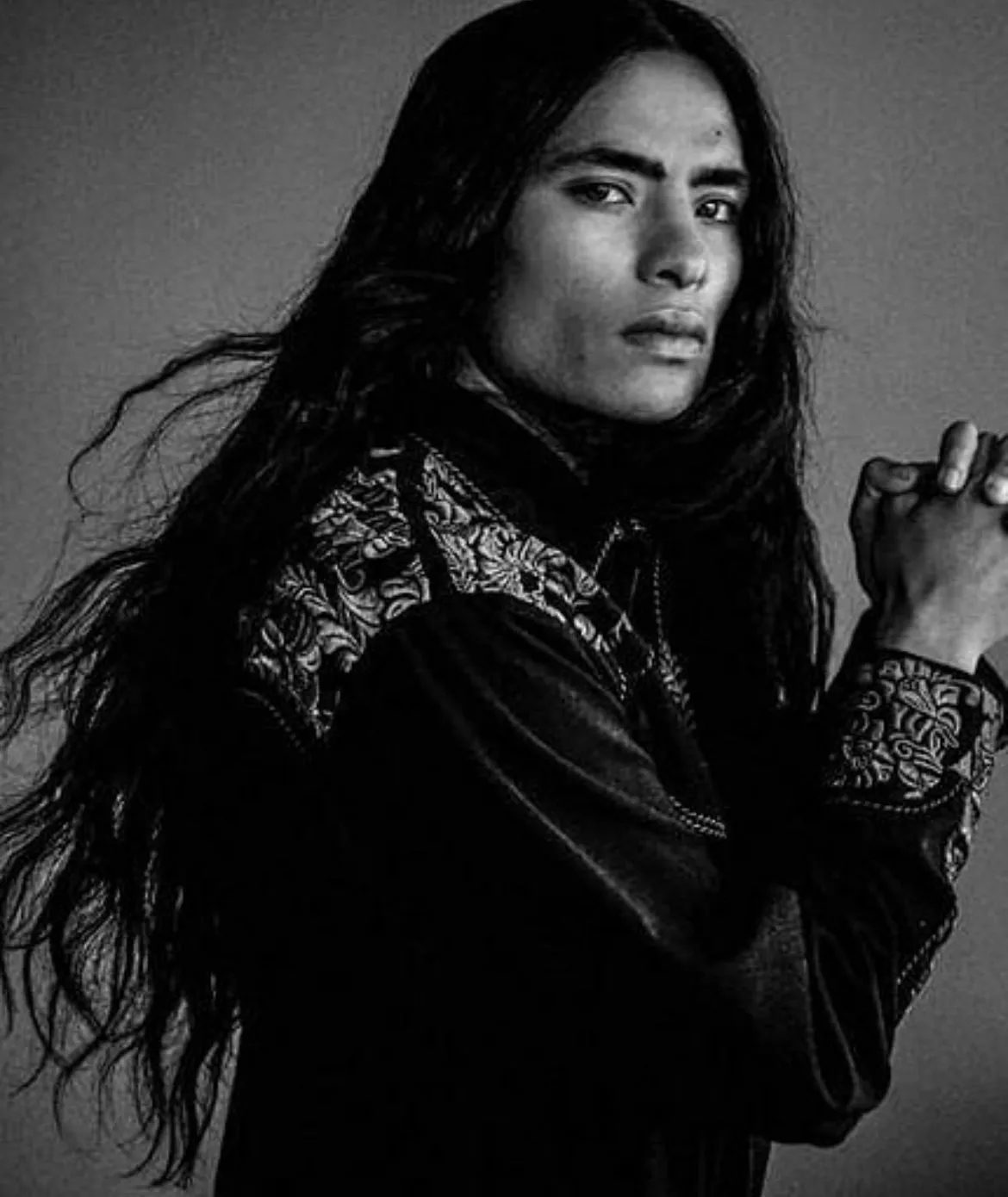 Man with long dark hair, wearing a dark shirt with embroidered detailing, hands clasped, looking at camera