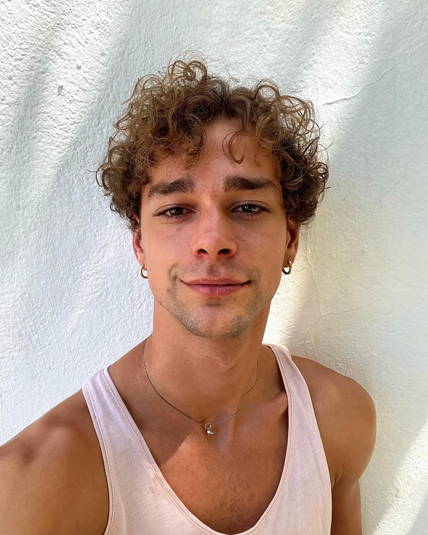 Close-up portrait of a young man with curly hair and small hoop earrings, wearing a light tank top and a thin necklace, standing against a textured white wall in natural daylight