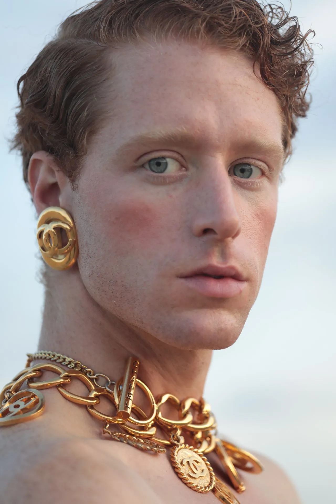 Man with red hair, gold jewelry, and light blush, looking towards the camera against a blurred blue background