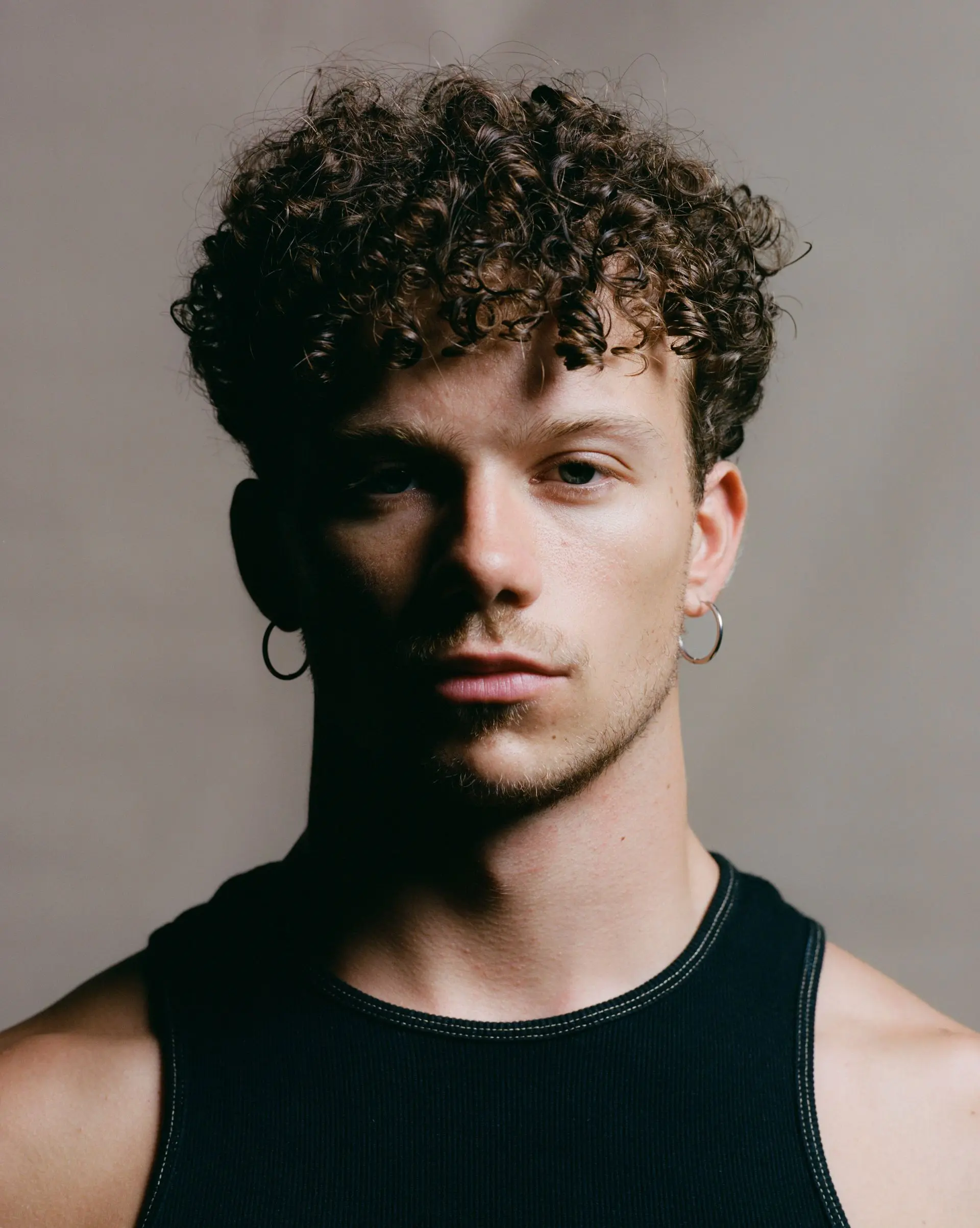 Studio portrait of a young man with short curly hair and hoop earrings, wearing a black sleeveless top, facing the camera against a neutral background with soft, dramatic lighting