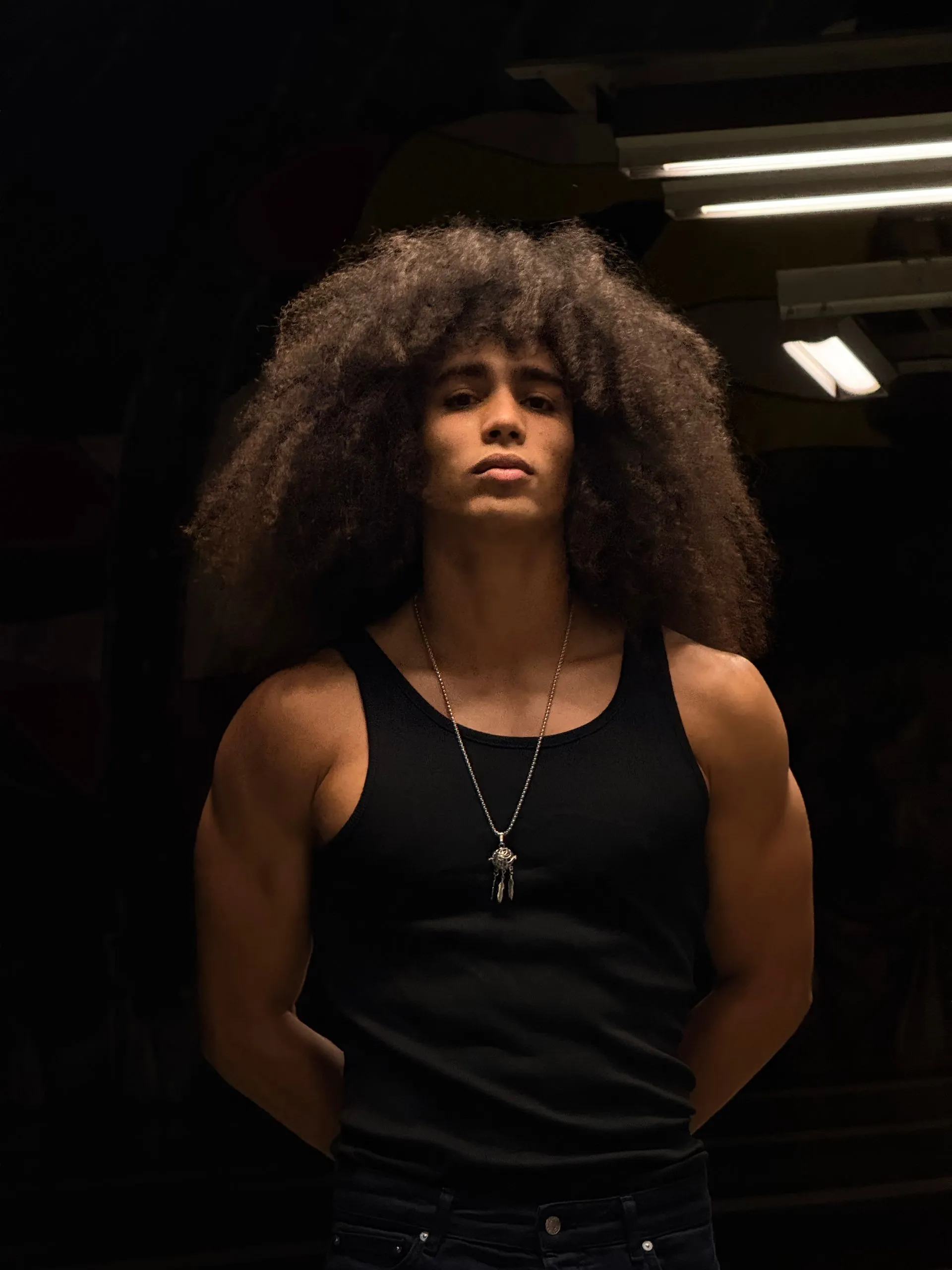 Man with large afro, arms crossed behind back, wearing black tank top. Standing in dim, dark area