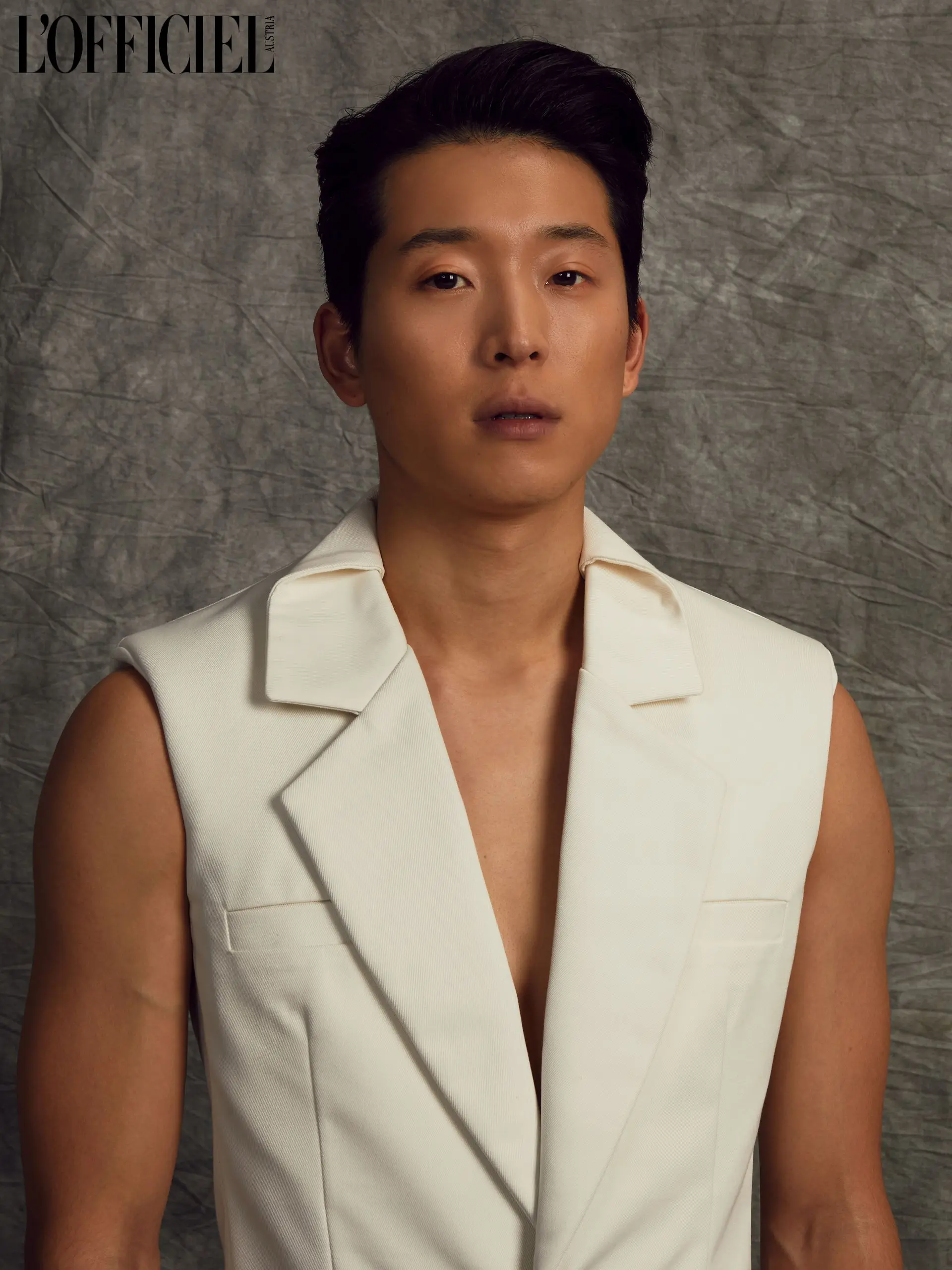 Man in white top, dark hair, neutral expression, against a grey backdrop