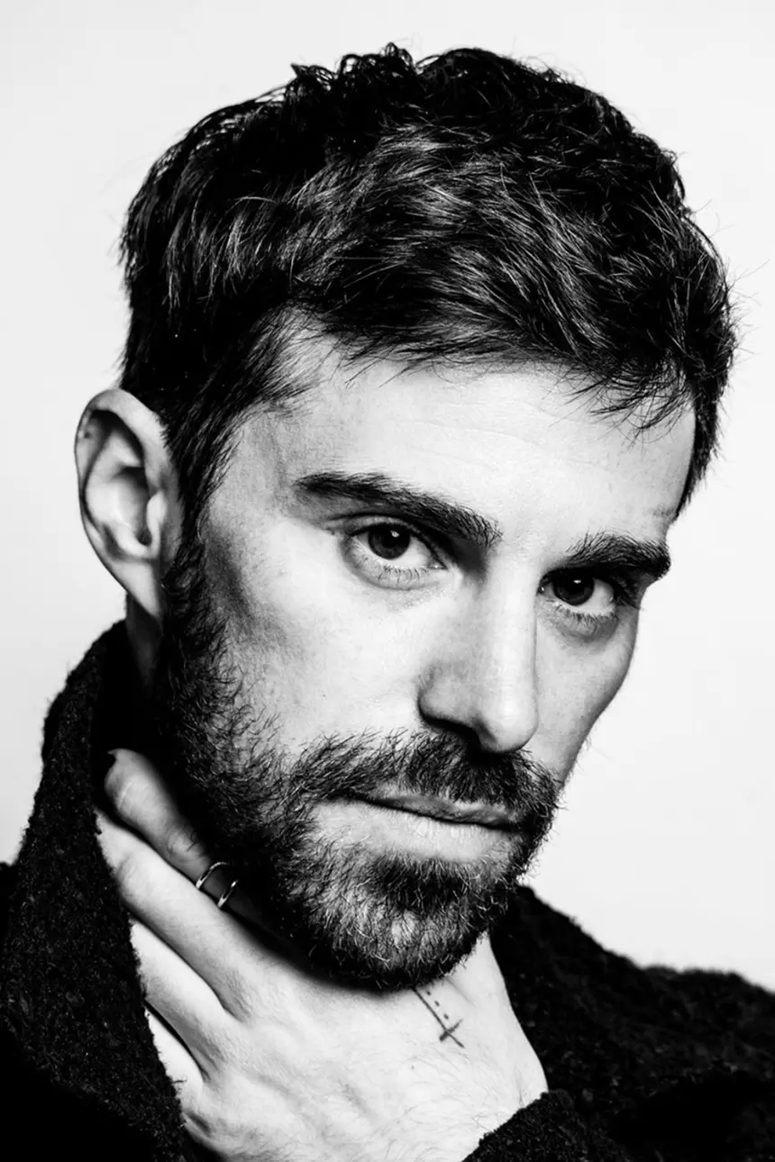 Man with dark hair and beard, looking intently at the camera, wearing a dark top, hands near his neck