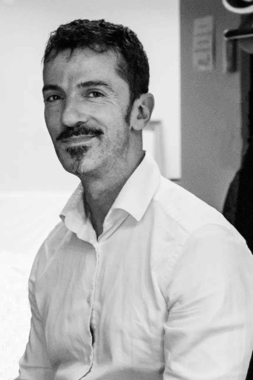 Man with dark hair and a goatee smiles, wearing a light-colored button-down shirt