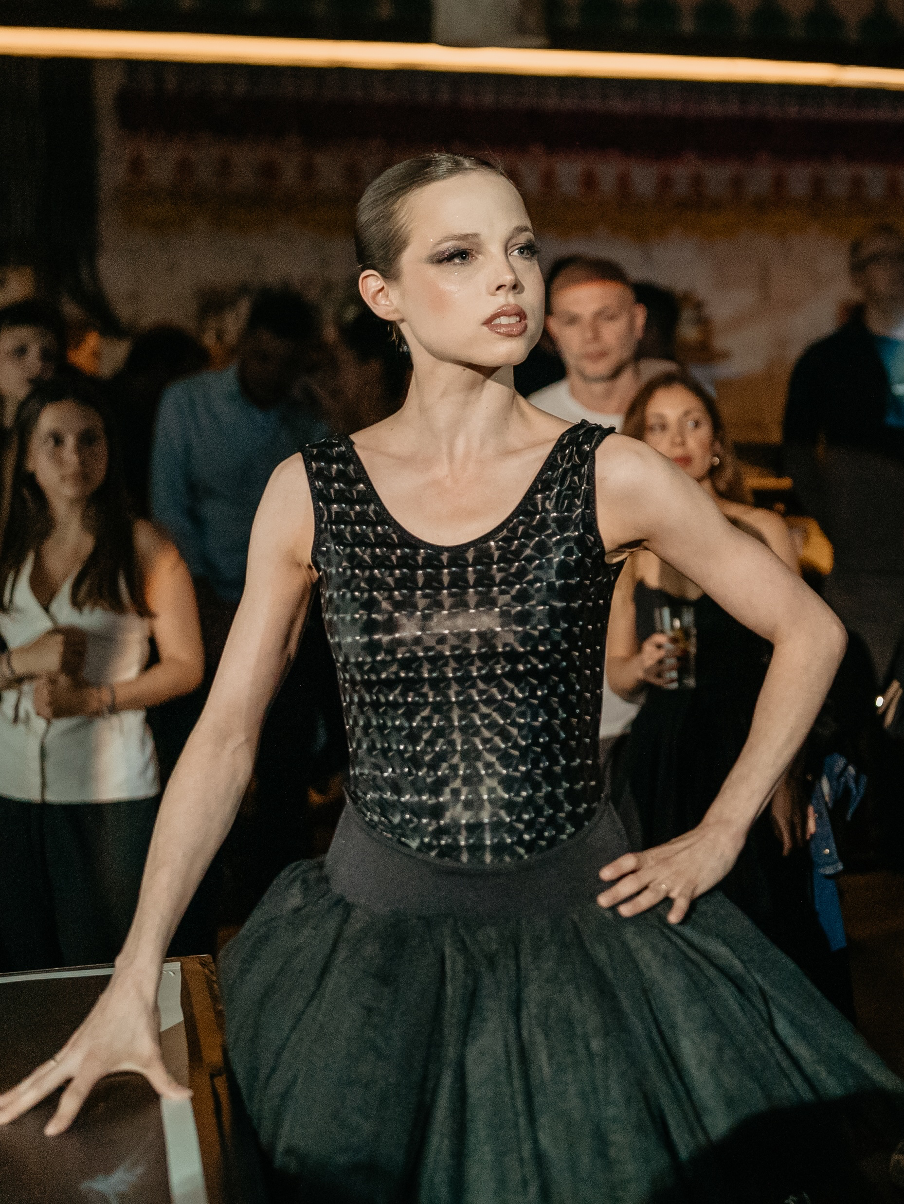 A portrait of a young woman with slicked-back hair wearing a sleeveless black, textured bodice and a full dark gray/black tulle skirt. She stands in a crowded indoor setting with one hand on her hip and the other resting on a table, looking slightly to the