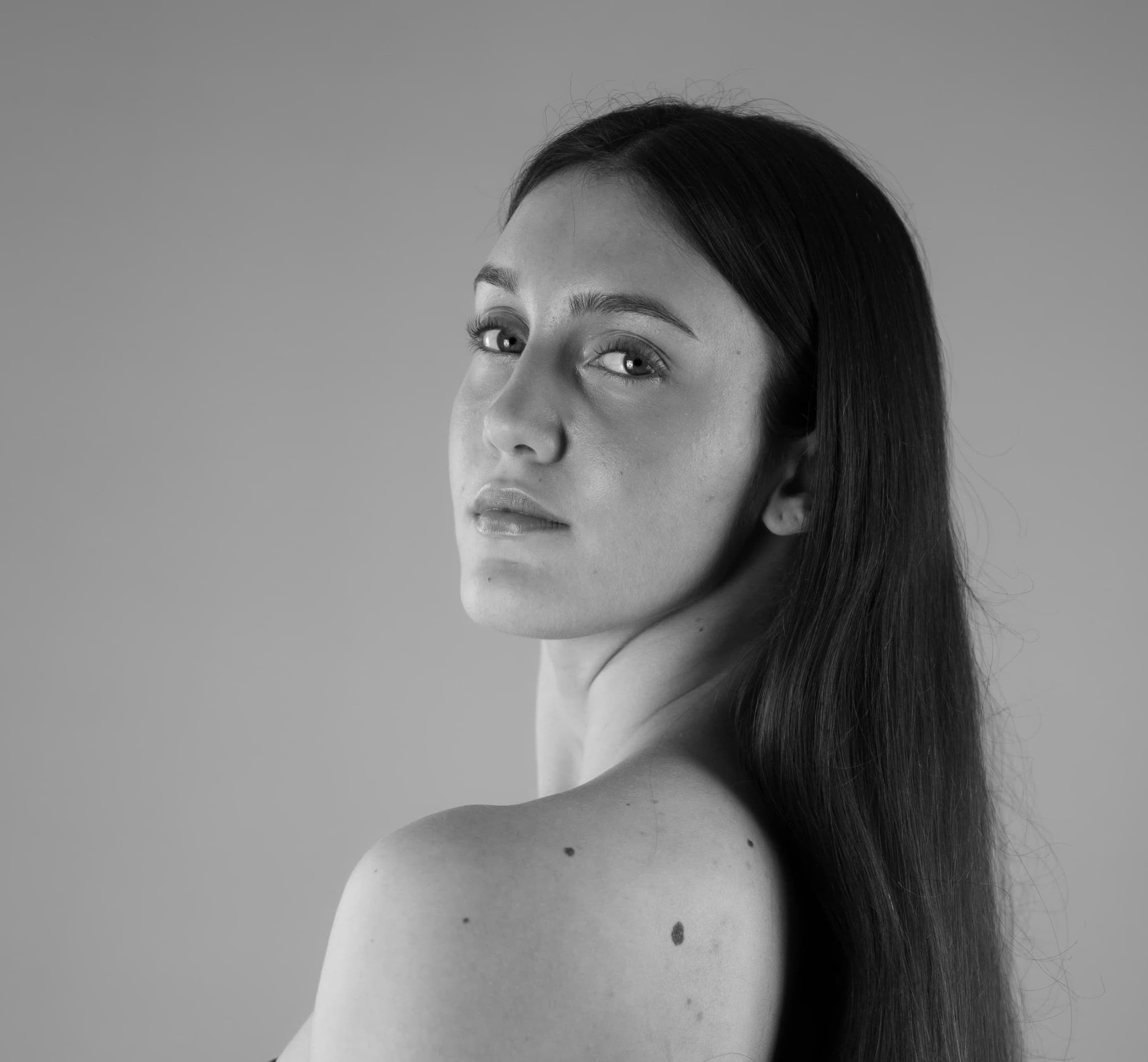 A studio black and white portrait of a young woman — an artist represented by a dance talent agency. She has long, straight dark hair and elegantly turns her head over a bare shoulder with a piercing gaze.