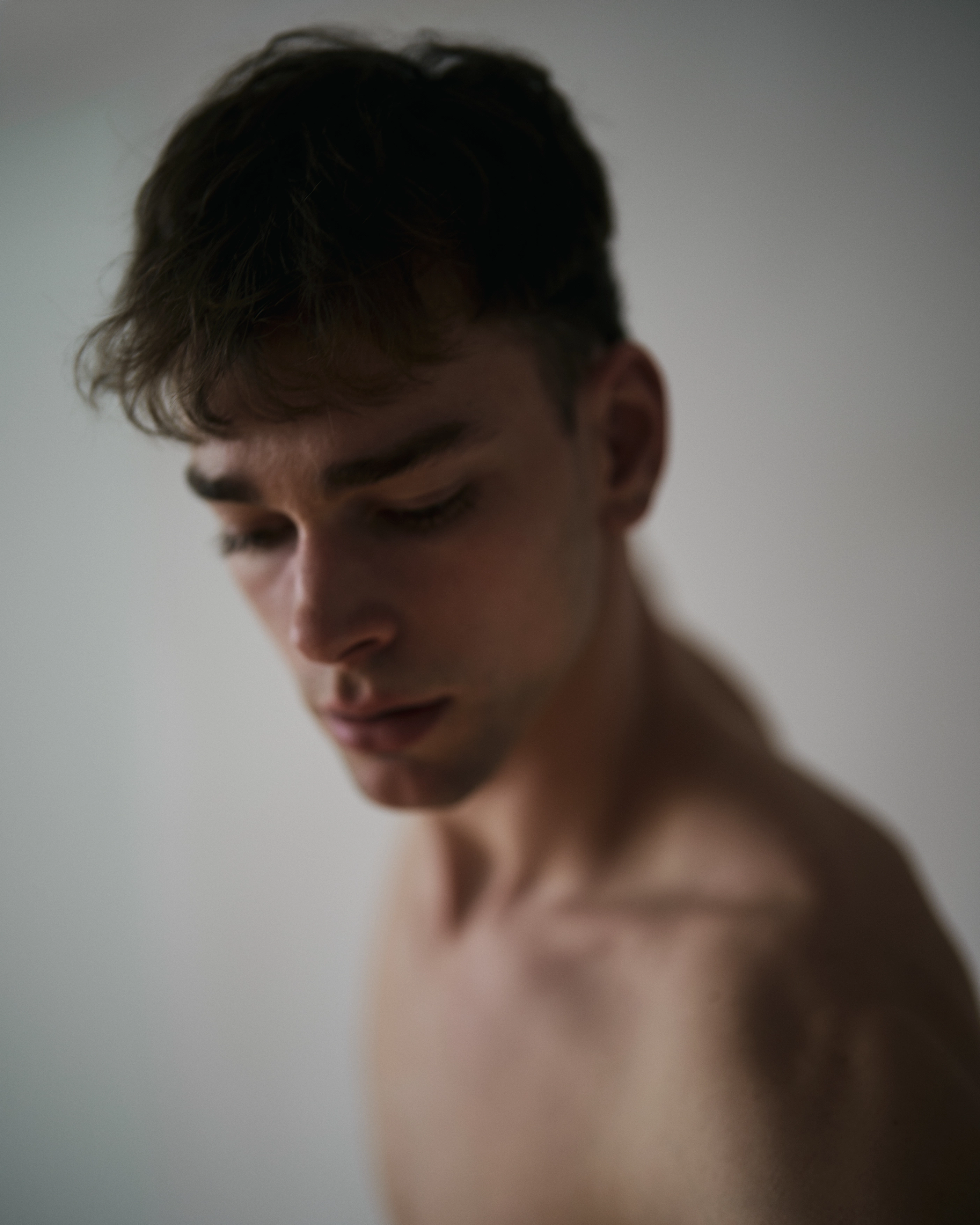 Nahaufnahme eines jungen Mannes mit lockigem Haar und kleinen Creolen, der ein helles Tanktop und eine dünne Halskette trägt und vor einer strukturierten weißen Wand im natürlichen Tageslicht steht