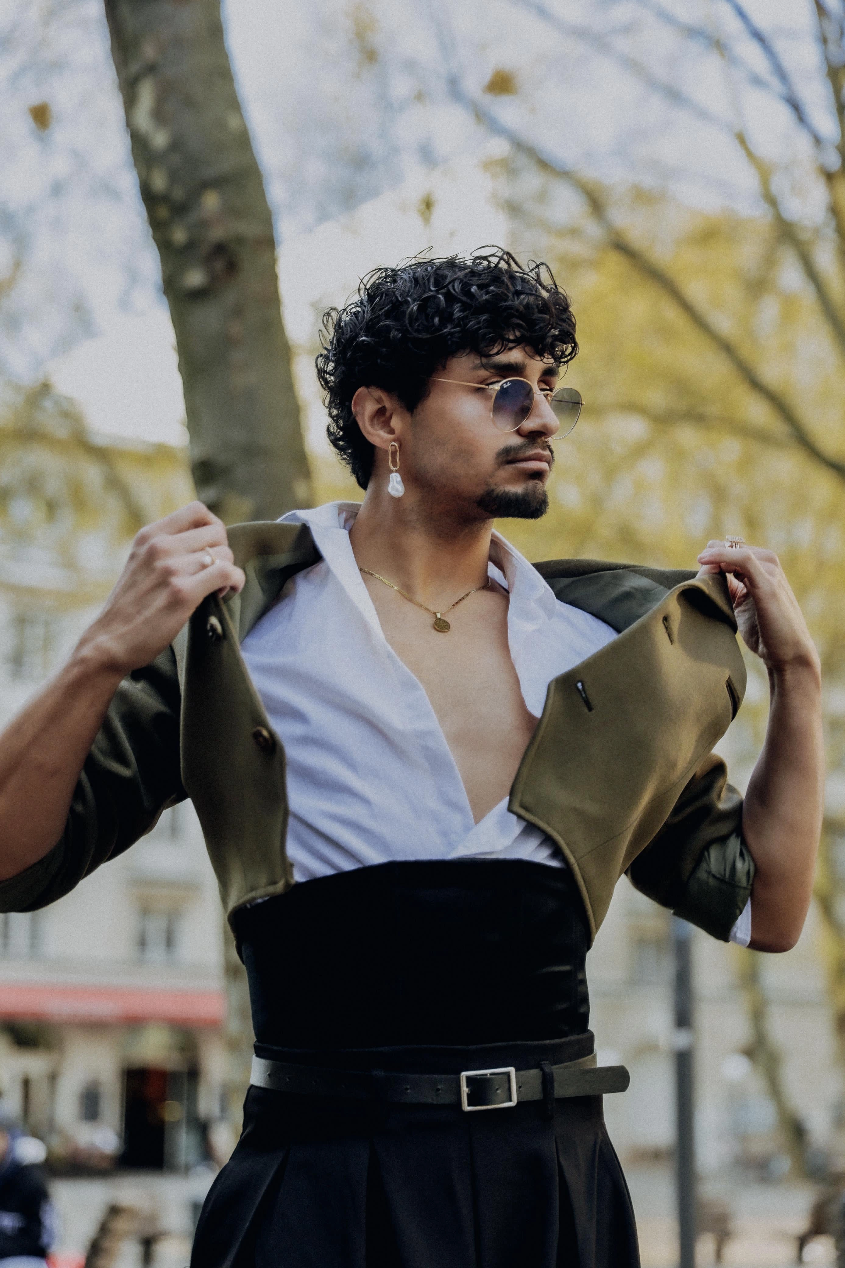 Portrait of a stylish man with curly hair and sunglasses, wearing an open green jacket over a white shirt, a black belt, and a black waist cincher, standing outdoors with trees in the background
