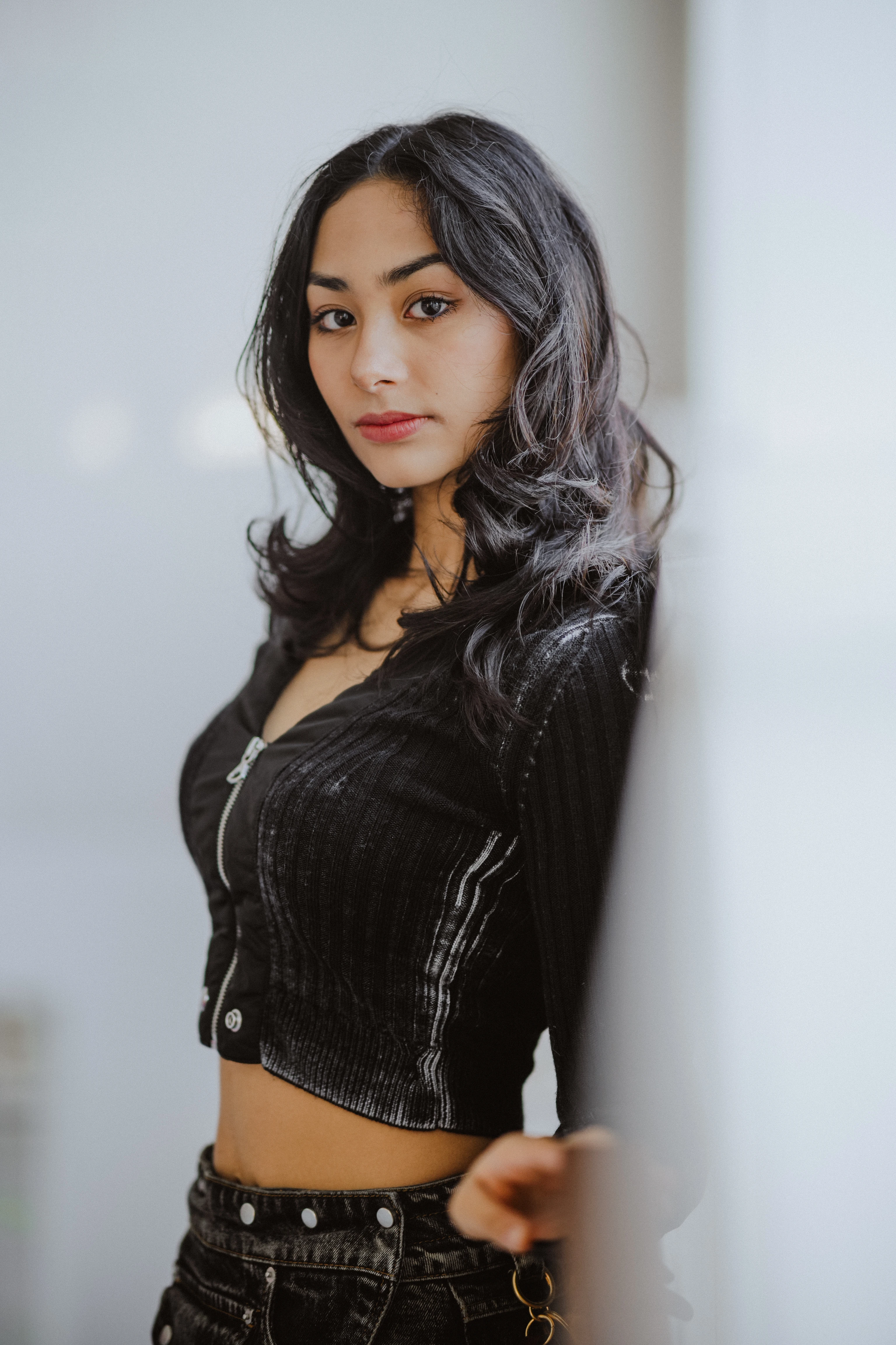 Porträt einer jungen Frau mit langen, dunklen Wellenhaaren, die sich gegen eine Wand lehnt, trägt eine schwarze kurze Strickjacke mit Reißverschluss und einen passenden schwarzen Gürtel, schaut in die Kamera.
