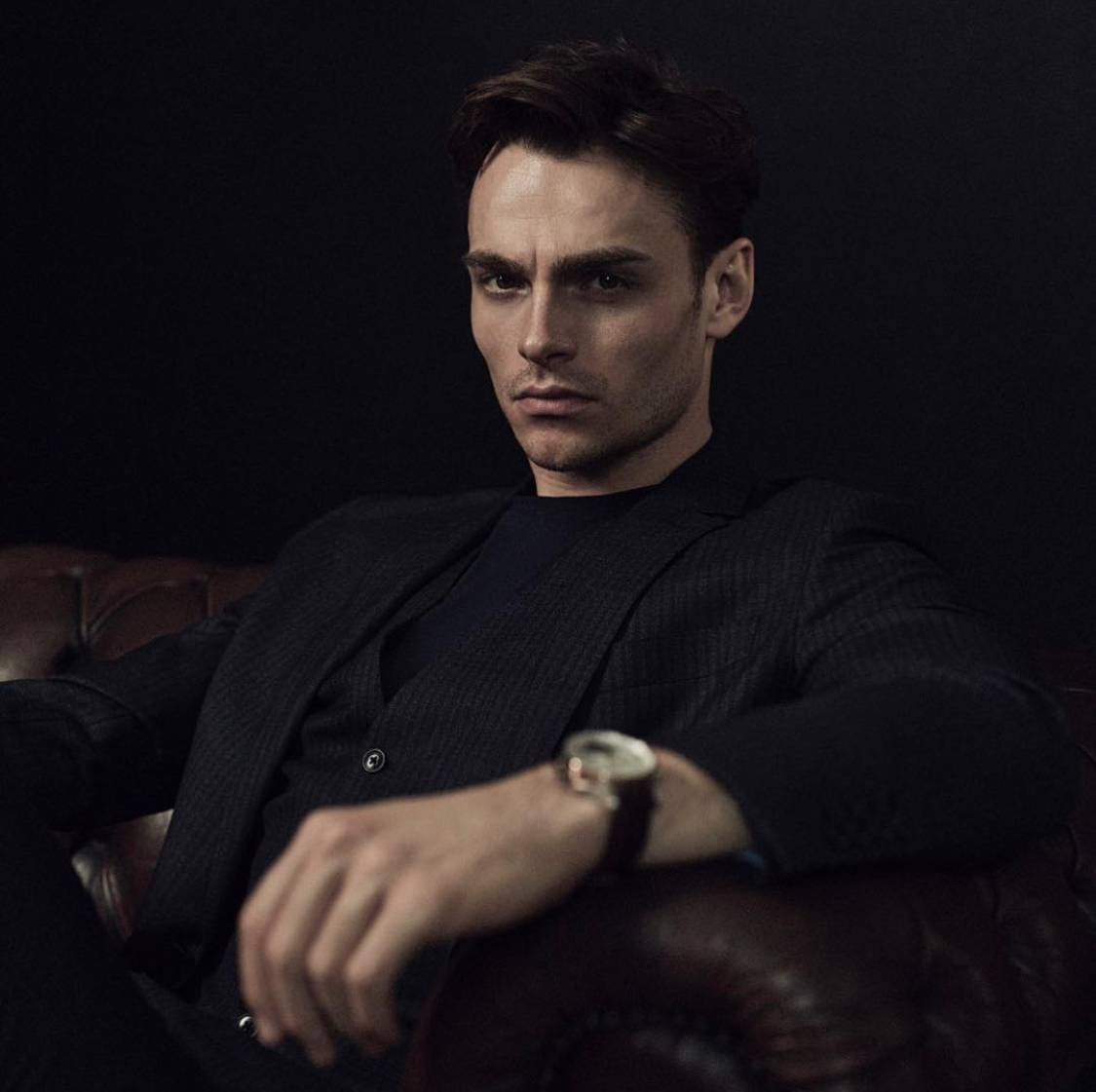 Portrait of a man with dark hair, wearing a black blazer and dark shirt, sitting on a brown leather sofa against a dark background, looking serious at the camera
