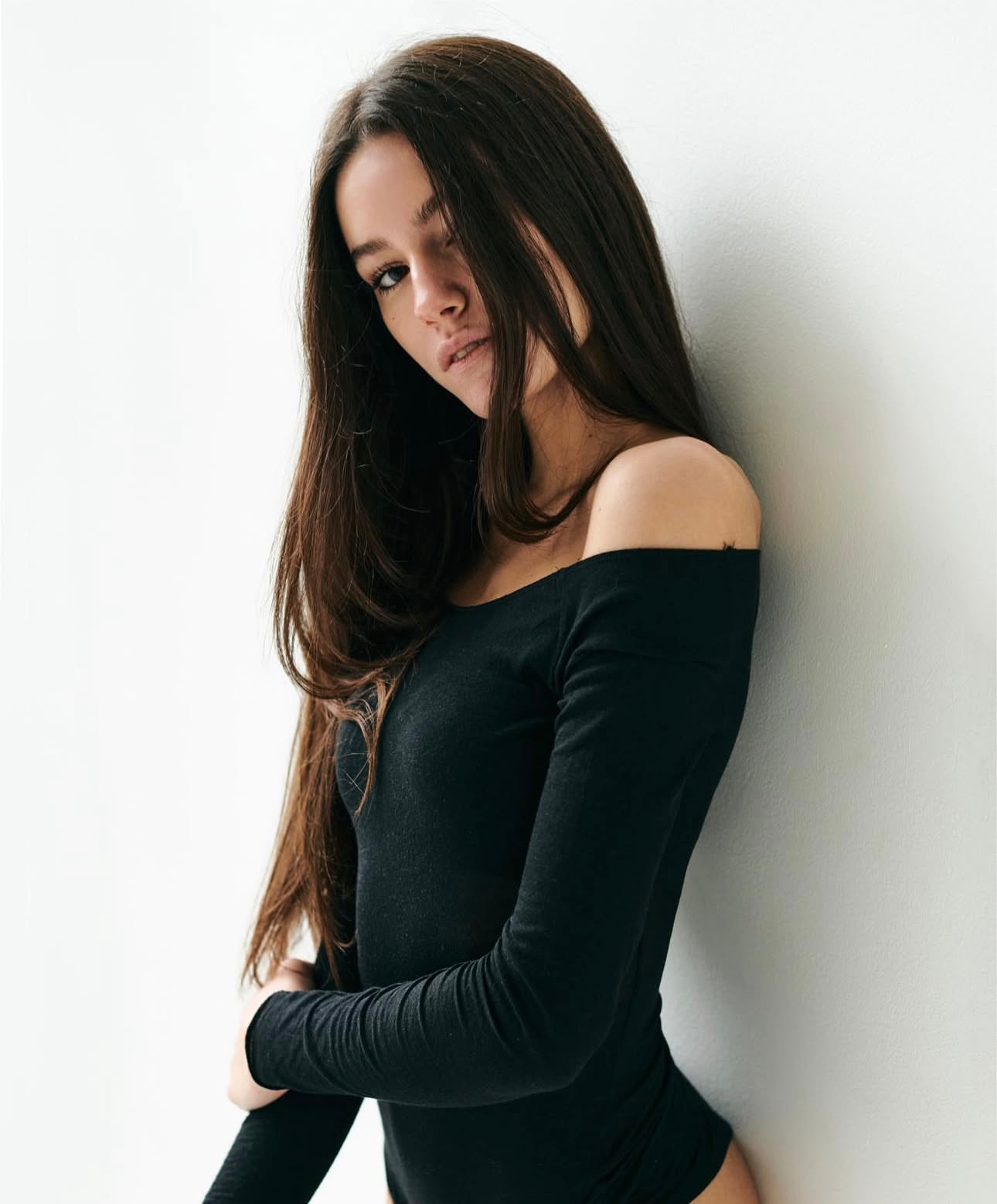 Junges weibliches Talent mit langen dunklen Haaren in einem schwarzen schulterfreien Body, an eine weiße Wand gelehnt. Selbstbewusste und elegante Pose.