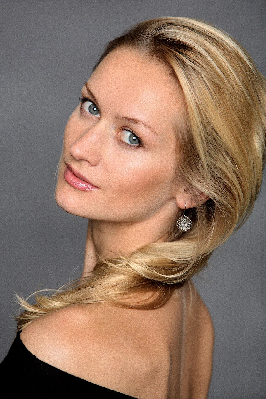 Young blonde female talent with blue eyes and elegant drop earrings, wearing a black off-shoulder top. Classic professional headshot against a grey background.