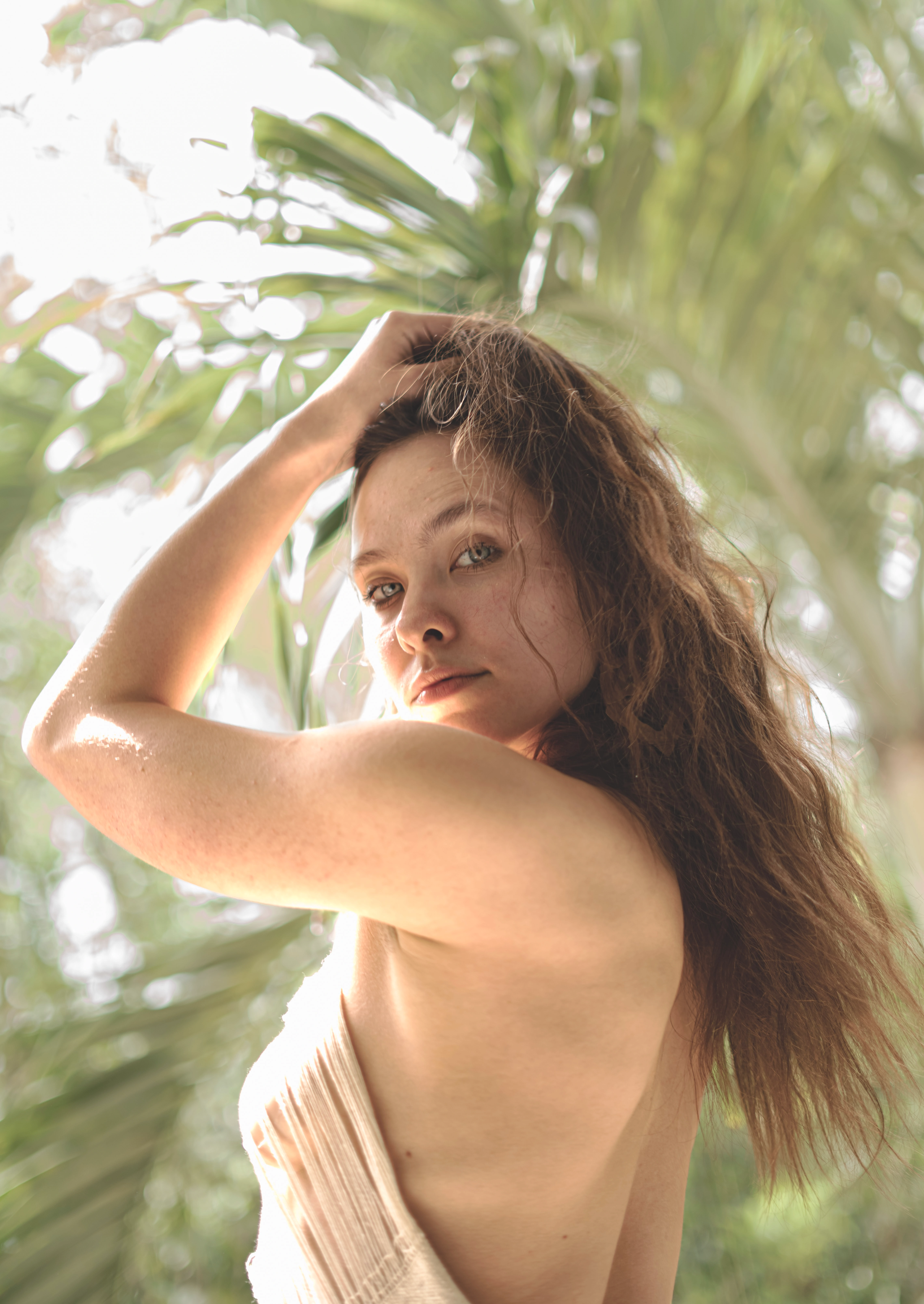 Junge Frau mit langen welligen rotbraunen Haaren posiert zwischen tropischen grünen Palmenblättern. Künstlerisches und ätherisches Außenportrait mit weichem natürlichem Licht.