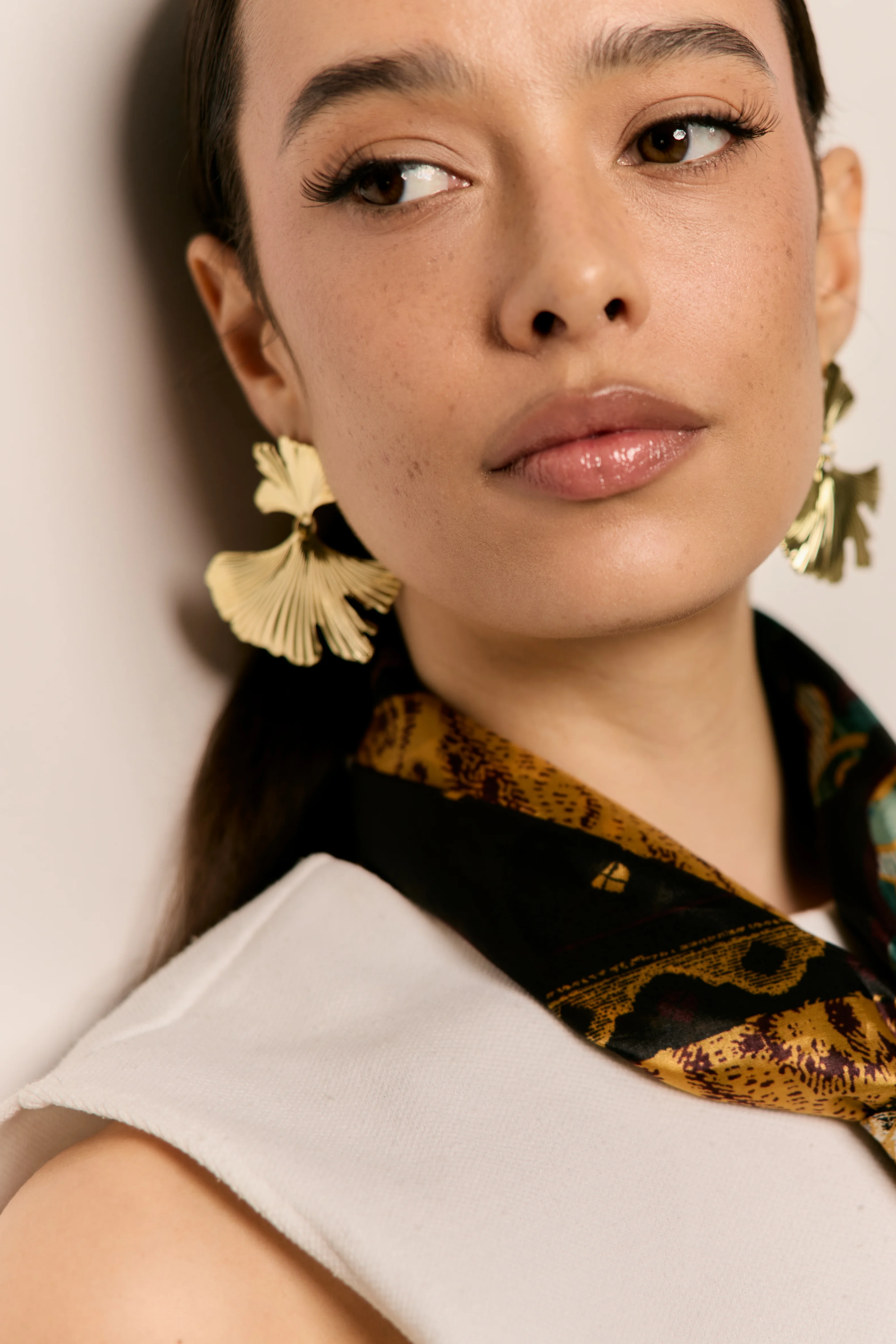 Nahaufnahme-Porträt einer jungen Frau mit dunklem Haar, goldenen Blumen-Ohrhängern, schwarz-goldenem Seidentuch und weißem ärmellosen Top. Markanter Winged Eyeliner, Nude-Lippen, heller Hintergrund. Elegante, editoriale Ästhetik.