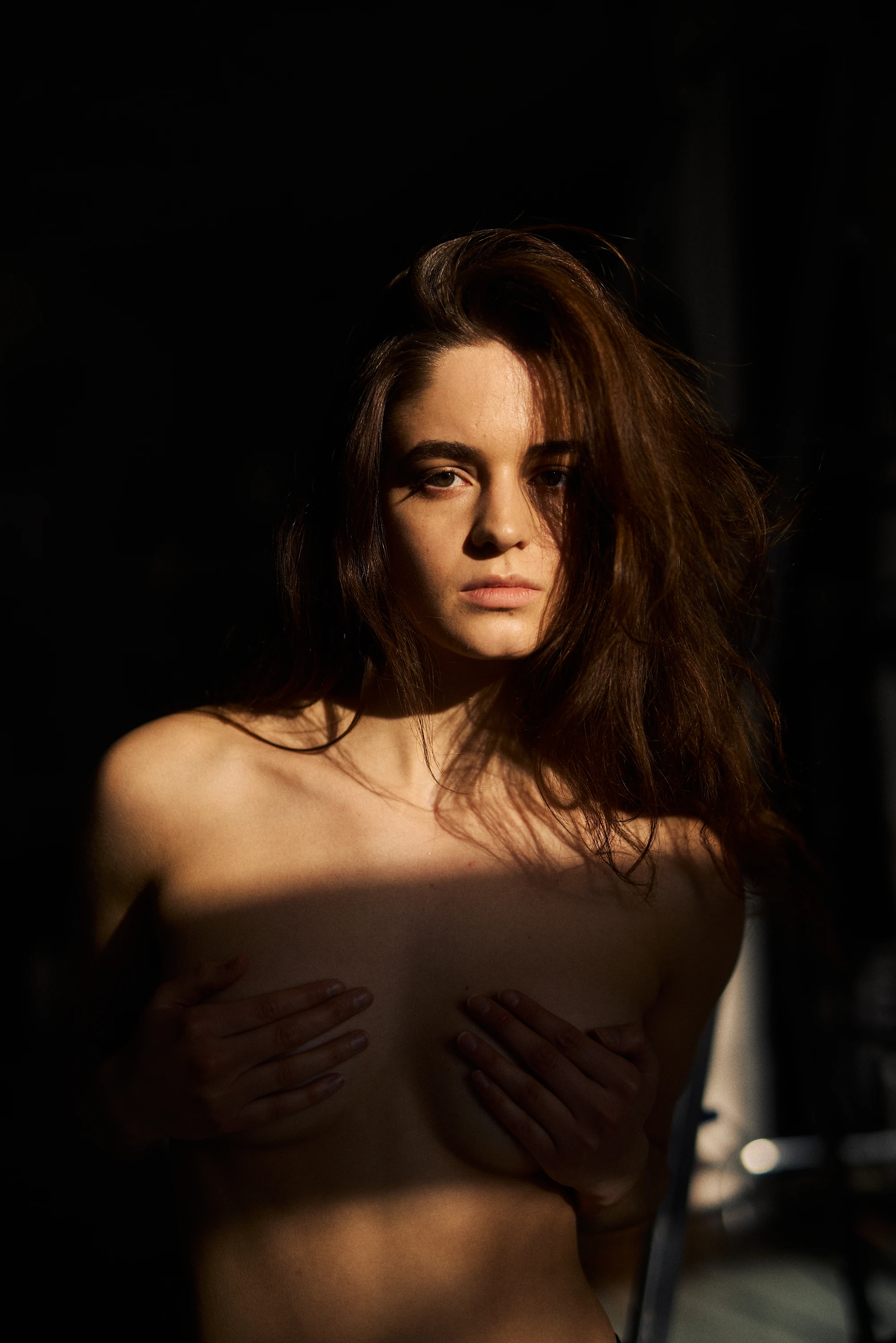 Artistic portrait of a young woman with long dark wavy hair, gazing downward with a serious expression. Dramatic shadows cross her bare shoulders and chest. She wears a delicate gold necklace. Dark background with strong chiaroscuro lighting creates a mood