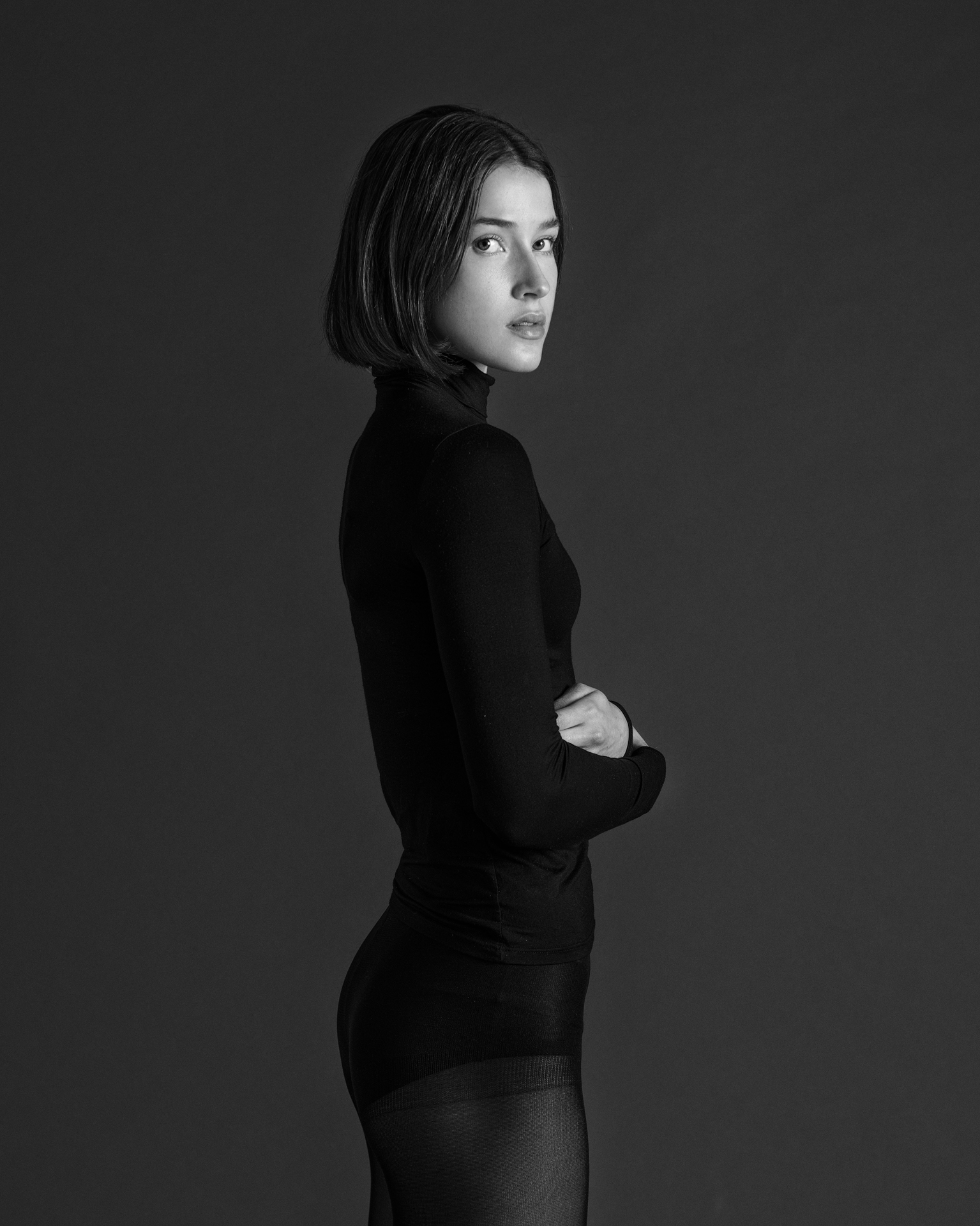 Schwarz-Weiß-Porträt einer jungen Frau mit kurzem Bob, schwarzem Rollkragenpullover, verschränkten Armen, die über die Schulter blickt – vor dunklem Hintergrund.
